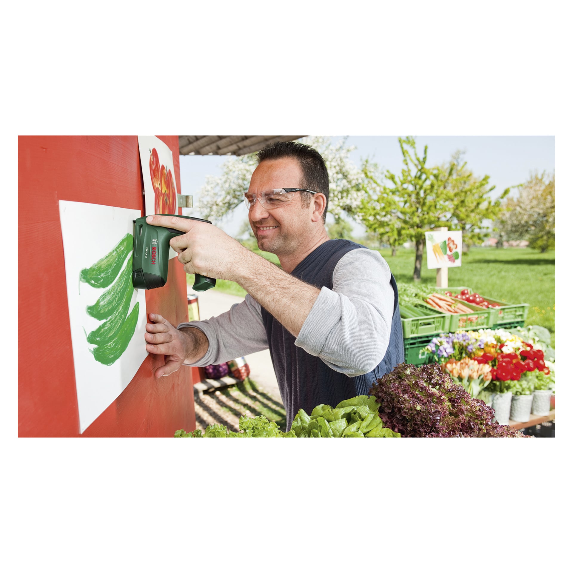 Ein Mann mit Schutzbrille benutzt eine Bosch PTK 3,6 LI batteriebetriebene Tackerpistole, um eine Zeichnung mit grünen Paprika an einer roten Wand zu befestigen. In der Nähe liegt frisches Gemüse auf einem Tisch im Sonnenlicht.