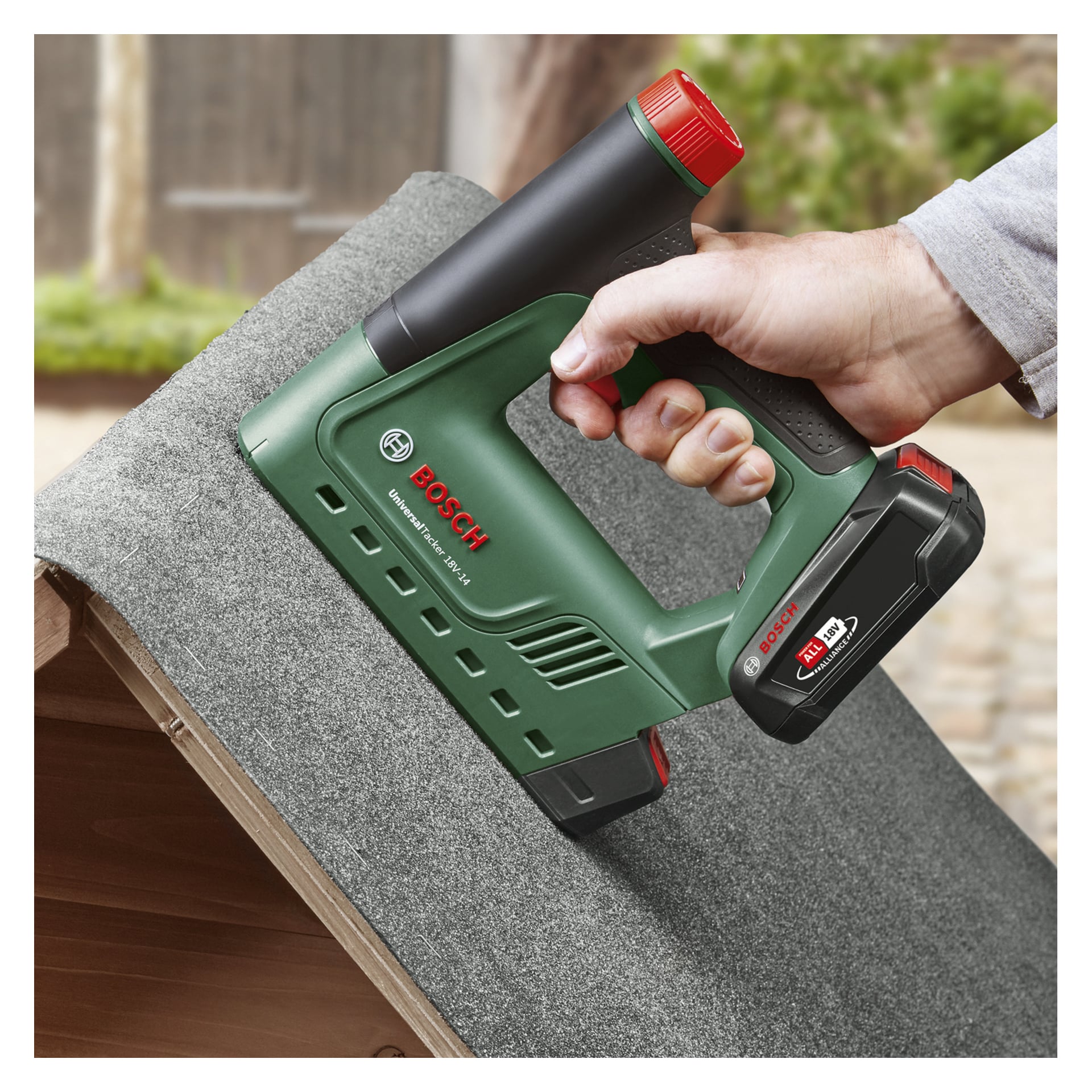 Jemand befestigt Dächerfilz mit einem grünen Bosch Power 4 All UniversalTacker 18V-14 Akkuhefter und einer Nagelpistole mit rotem Abzug und Batterie, die das Werkzeug fest am Griff hält, an einem Holzrahmen im Außenbereich.