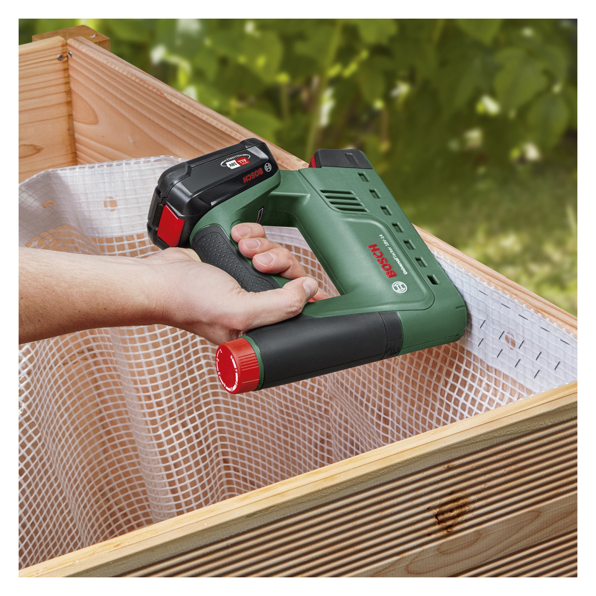 Jemand bedient eine grün-schwarze Bosch Power 4 All UniversalTacker 18V-14 batteriebetriebene Klammer-/Nagelpistole, die weißes Gitter an einem Holzrahmen befestigt. Der Hintergrund ist mit sichtbarem grünem Laub verschwommen.