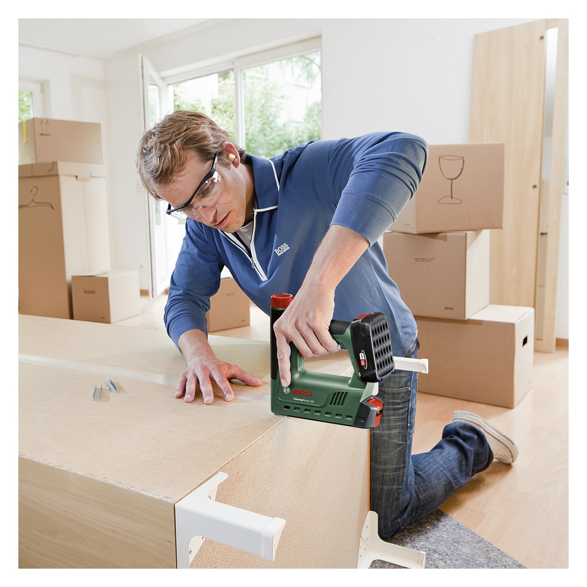 Eine Person mit Schutzbrille bedient eine Bosch Power 4 All Battery-betriebene Tacker-/Nagelpistole UniversalTacker 18V-14 und baut Holzmöbel in einem sonnenbeschienenen Raum zusammen, der mit Umzugskartons übersät ist und durch große Fenster erhellt wird.
