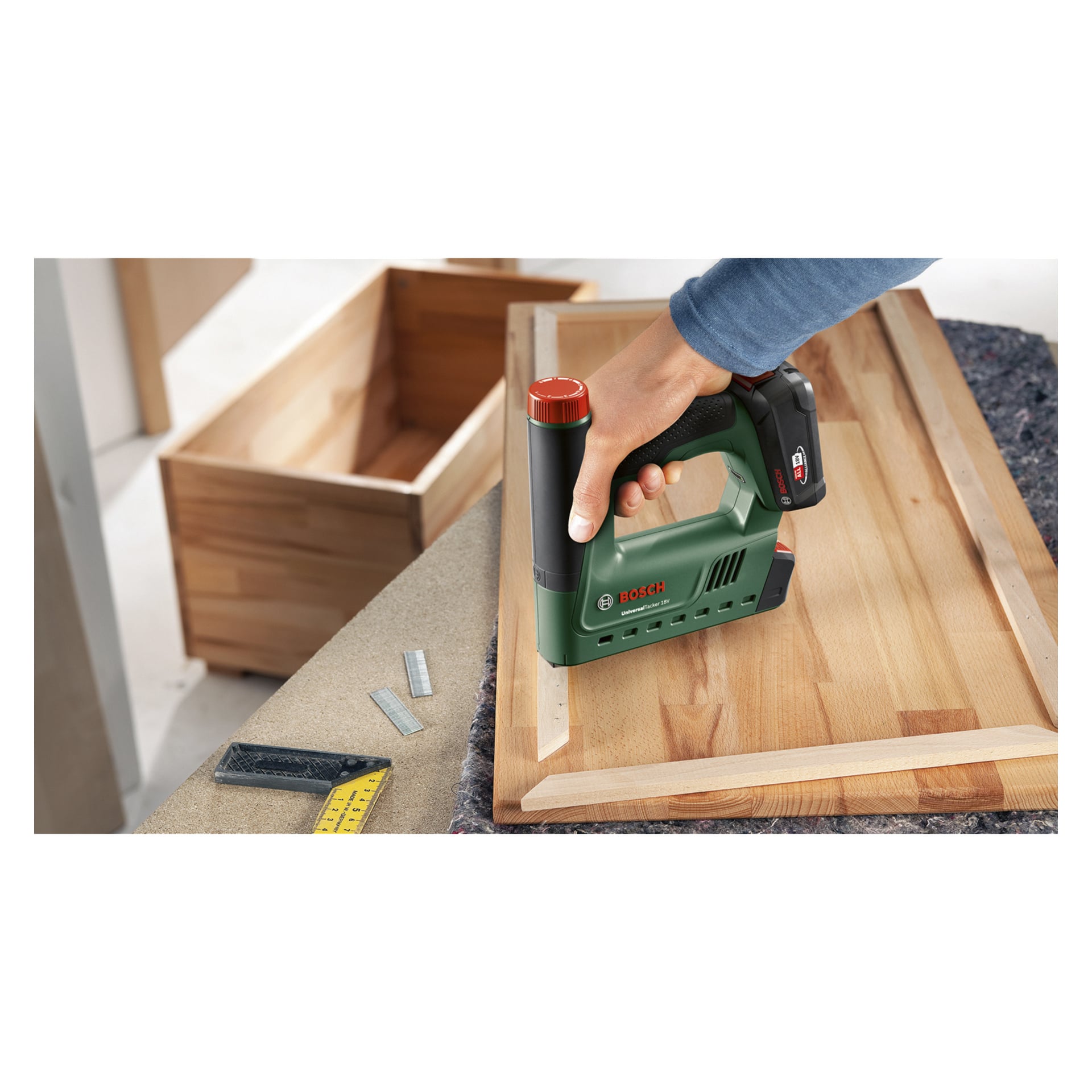 Jemand bedient eine Bosch Power 4 All UniversalTacker 18V-14 batteriebetriebene Klammer und Nagelpistole auf einem Holzrahmen. Auf der mit Stoff bedeckten Werkbank befinden sich lose Klammern, ein gelbes Maßlineal und eine Holzschublade.