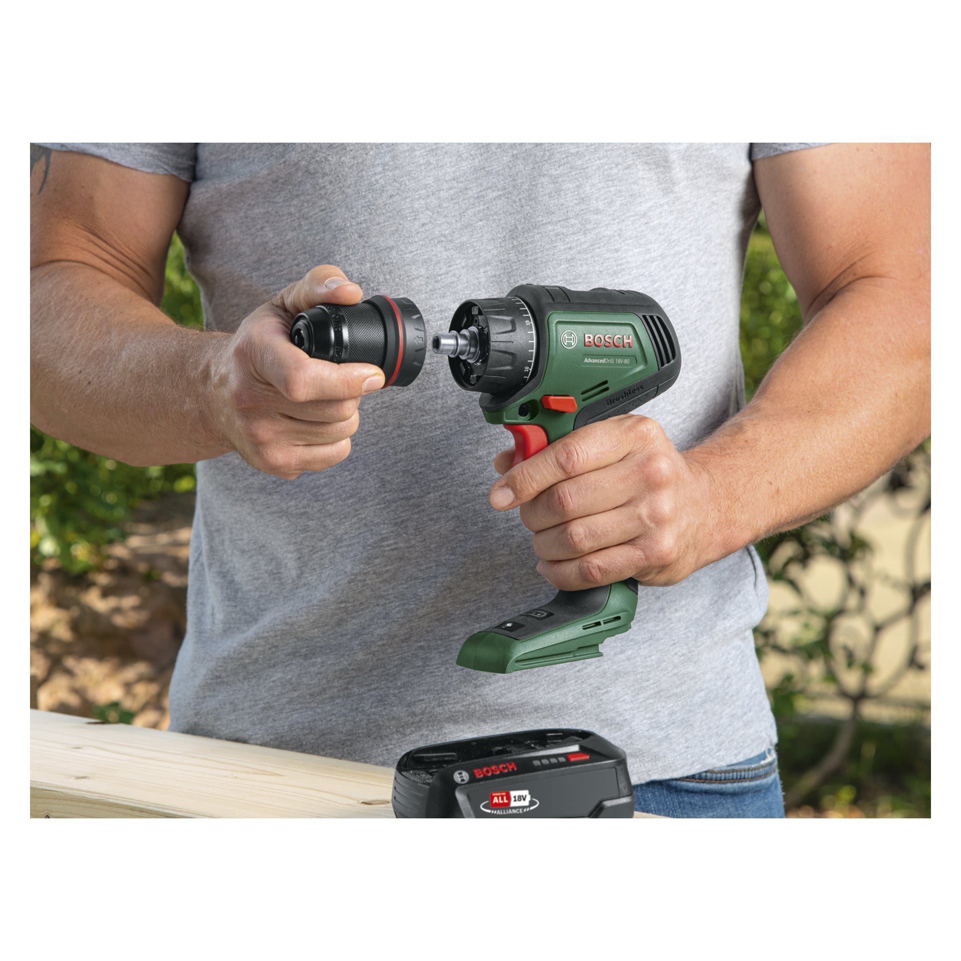 Harmaaseen paitaan pukeutunut henkilö on ulkona, pitäen Bosch Power 4 All Brushless AdvancedDrill 18V-80 -ruuvimeisseliä toisessa kädessään ja kiinnittäen mustaa poranterän pidikettä toisella. Lähellä olevalla puupinnalla on Boschin akkupaketti.