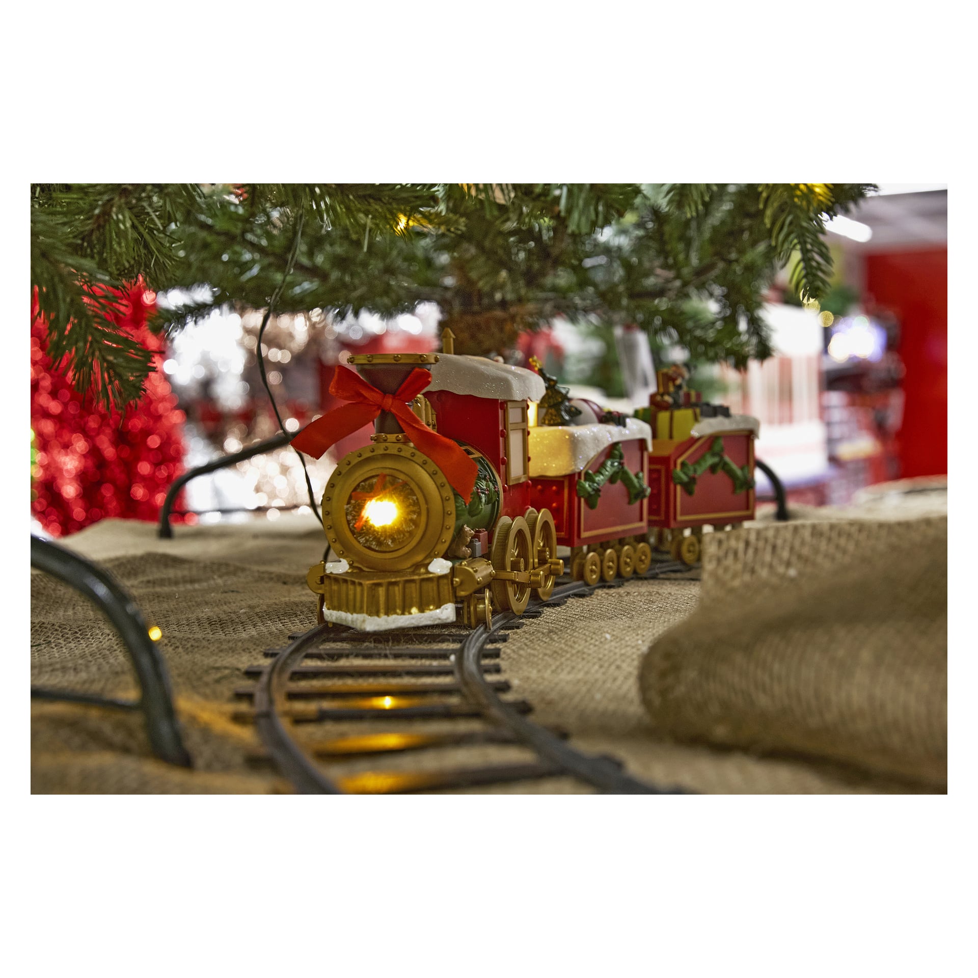 Ein festlicher EKVIP-Weihnachtszug, dekoriert in roten und goldenen Farben, Kränzen und einem Band, dessen Scheinwerfer hell leuchtet, steht auf Schienen unter dem Weihnachtsbaum, wobei Jute um den Sockel gewickelt ist.