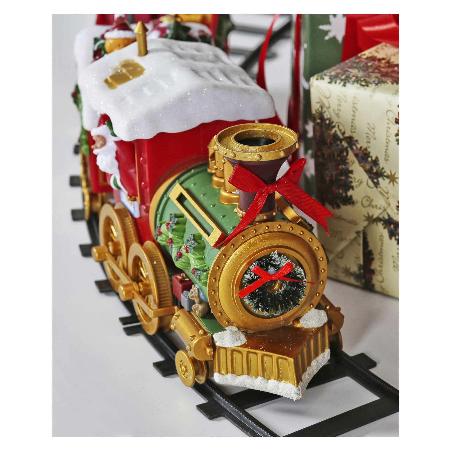 Ein festlicher EKVIP Weihnachtszug, geschmückt in Rot, Grün und Gold mit schneebedeckten Akzenten, einer leuchtend roten Schleife und einem Kranz vorne, ruht auf schwarzen Schienen neben einem verpackten Weihnachtsgeschenk.