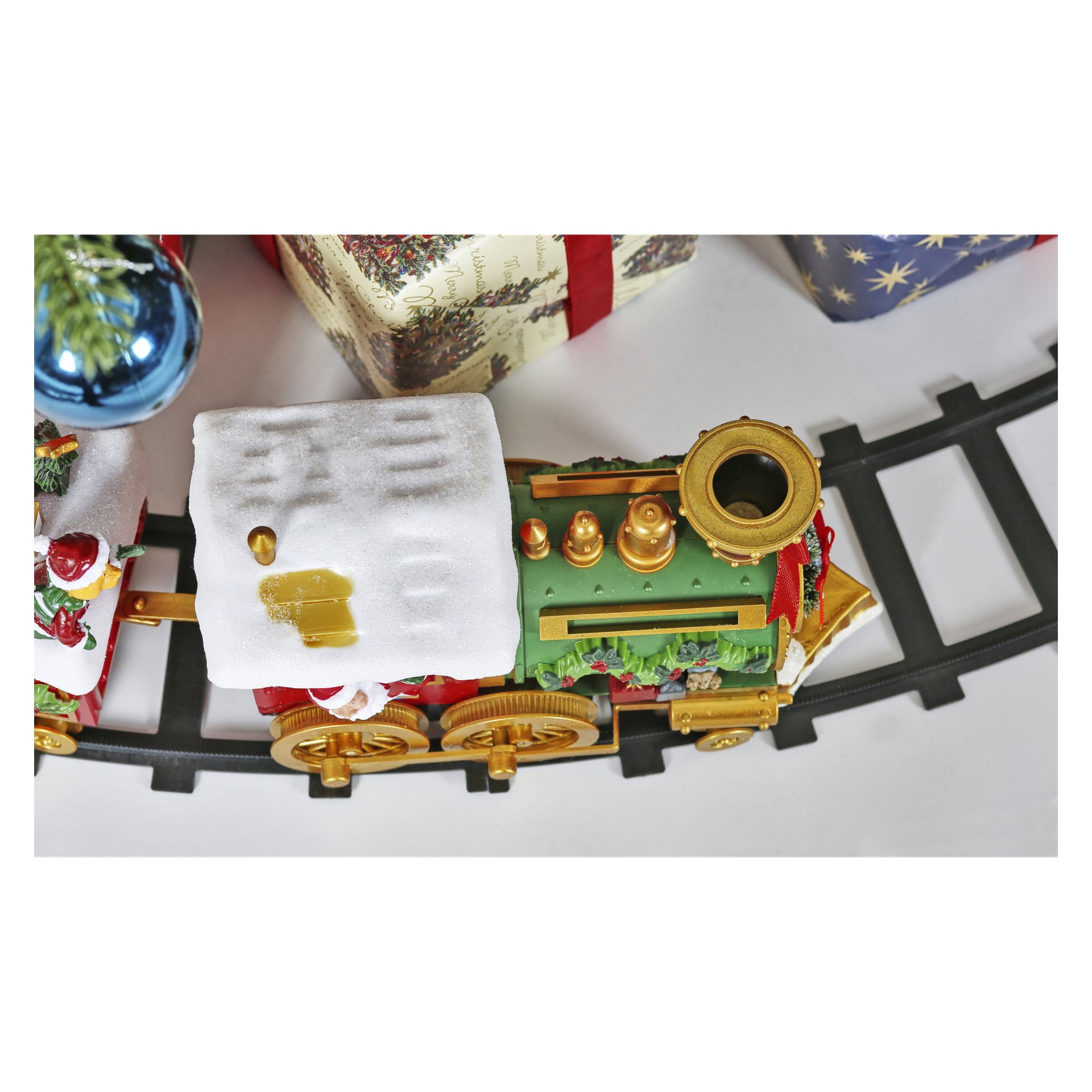 Eine festliche EKVIP Weihnachtslokomotive, mit bunten Akzenten dekoriert und mit weißen, schneeartigen Details bestreut, steht auf einer geschwungenen Strecke. Teilweise dahinter sind verpackte Geschenke und ein blauer Schmuck zu sehen.