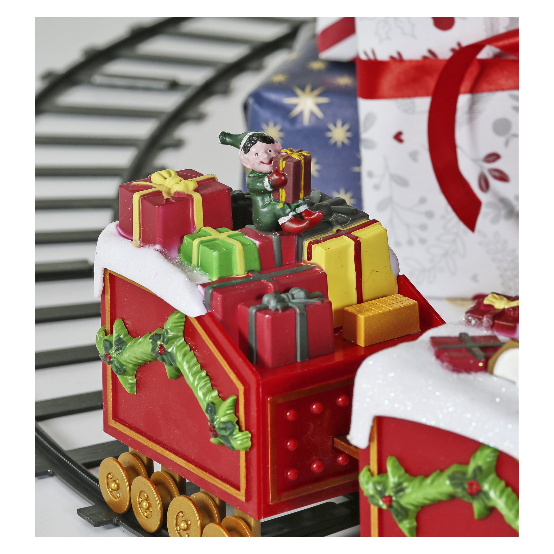 Ein festlicher EKVIP-Weihnachtszug zeigt ein Spielzeugauto, das mit grüner Girlande geschmückt ist, mit bunten Geschenkboxen und einem fröhlichen kleinen Elf mit einem Geschenk ausgestattet ist. Im Hintergrund erscheinen weitere verpackte Geschenke und Stücke von Eisenbahnschienen.