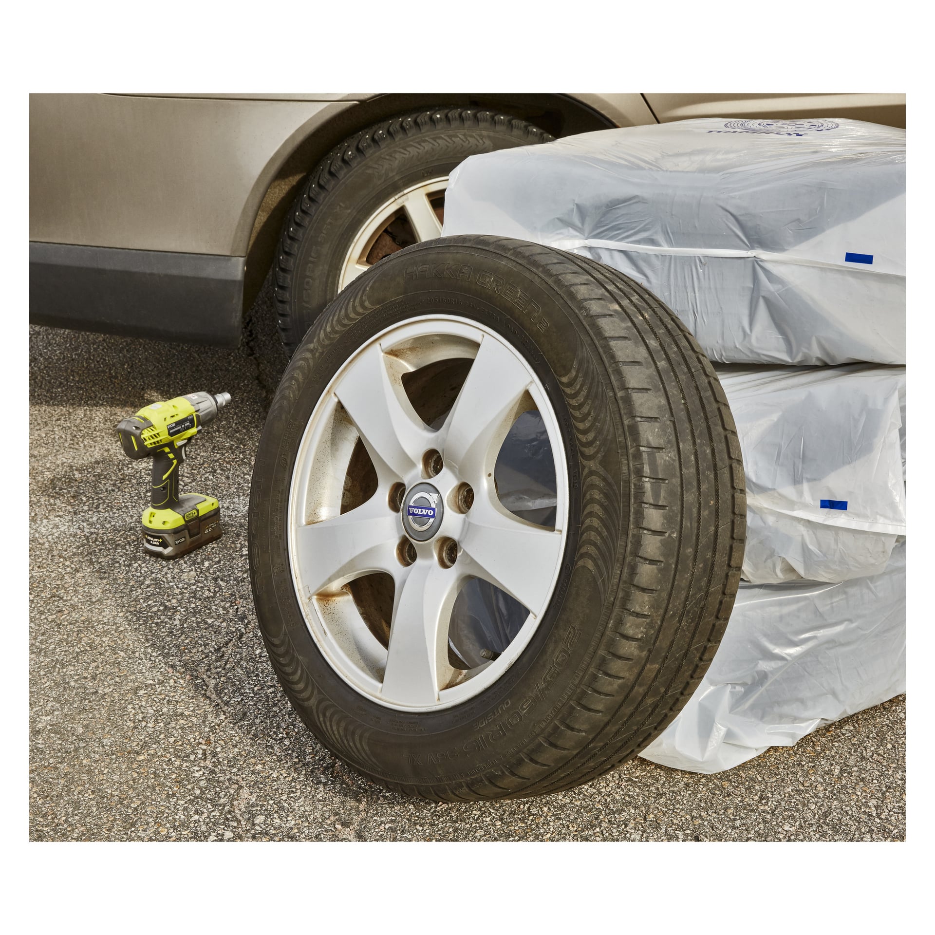 Auton rengas, jossa on hopeinen Volvo-vanne, seisoo pystyssä asfaltilla pinottujen renkaiden vieressä, jotka on kääritty pussiin, joissa lukee "Tyre bags 4 pack by Hamron." Paikalla on johdeton porakone ja osa toista autoa.