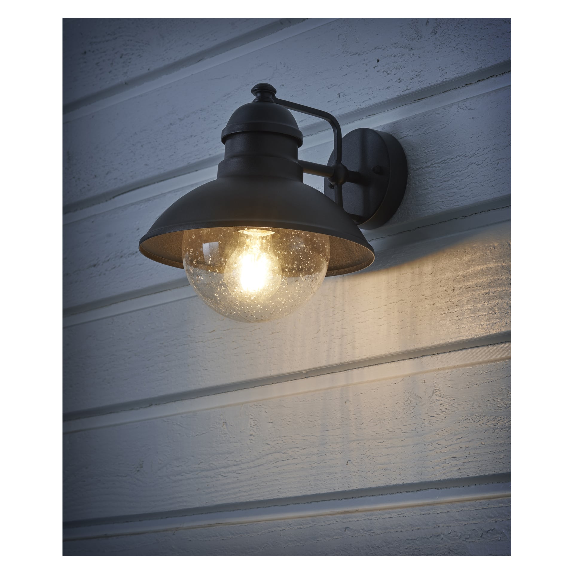 An einer hellblauen Holzwand montiert, hat die EKVIP-Wandlampe einen schwarzen Metallrahmen mit einer klaren Glaskugel. Die leuchtende Glühbirne darin wirft ein gemütliches, warmes Licht.