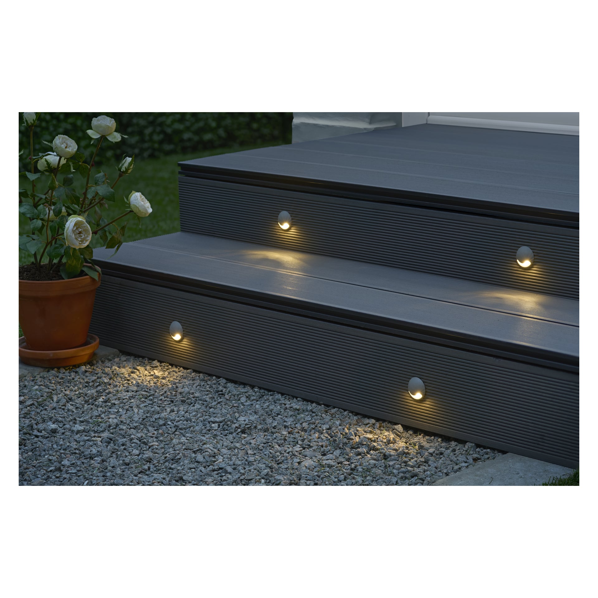 Bei Einbruch der Dämmerung werden zwei graue Deckstufen von eingebauten runden LED-Lichtern aus dem Anslut Steplights Connectable System 4-Pack beleuchtet. Eine Topfpflanze mit weißen Blüten steht links von den Treppen, die von einem Kiesweg nach oben führen. Im Hintergrund erscheinen Gras und eine Hecke.