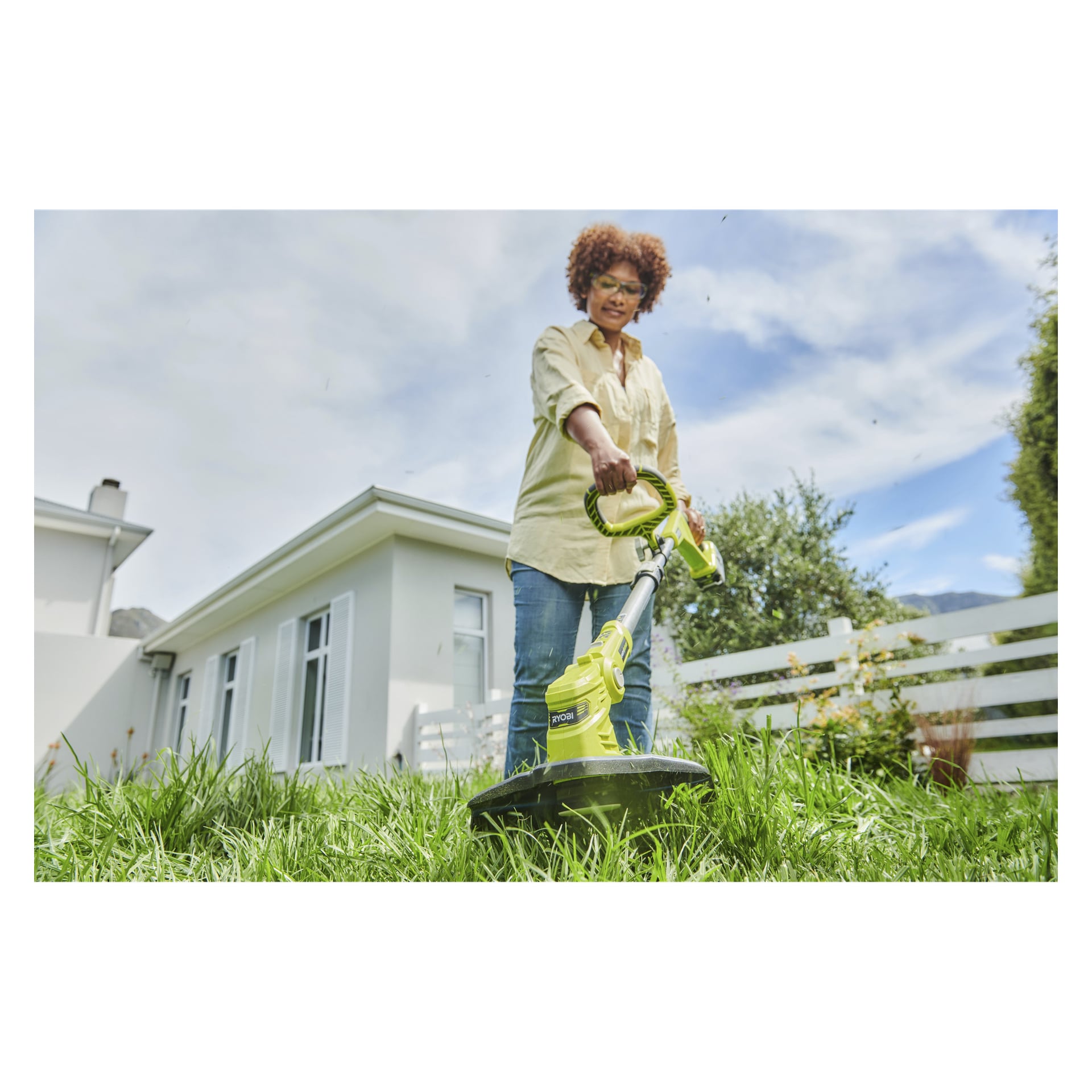 Någon klipper gräsmattan i en bostadsträdgård med en Ryobi One+ batteridriven RLT183240F grästrimmer, med ett vitt hus och staket synligt bakom sig en solig dag.