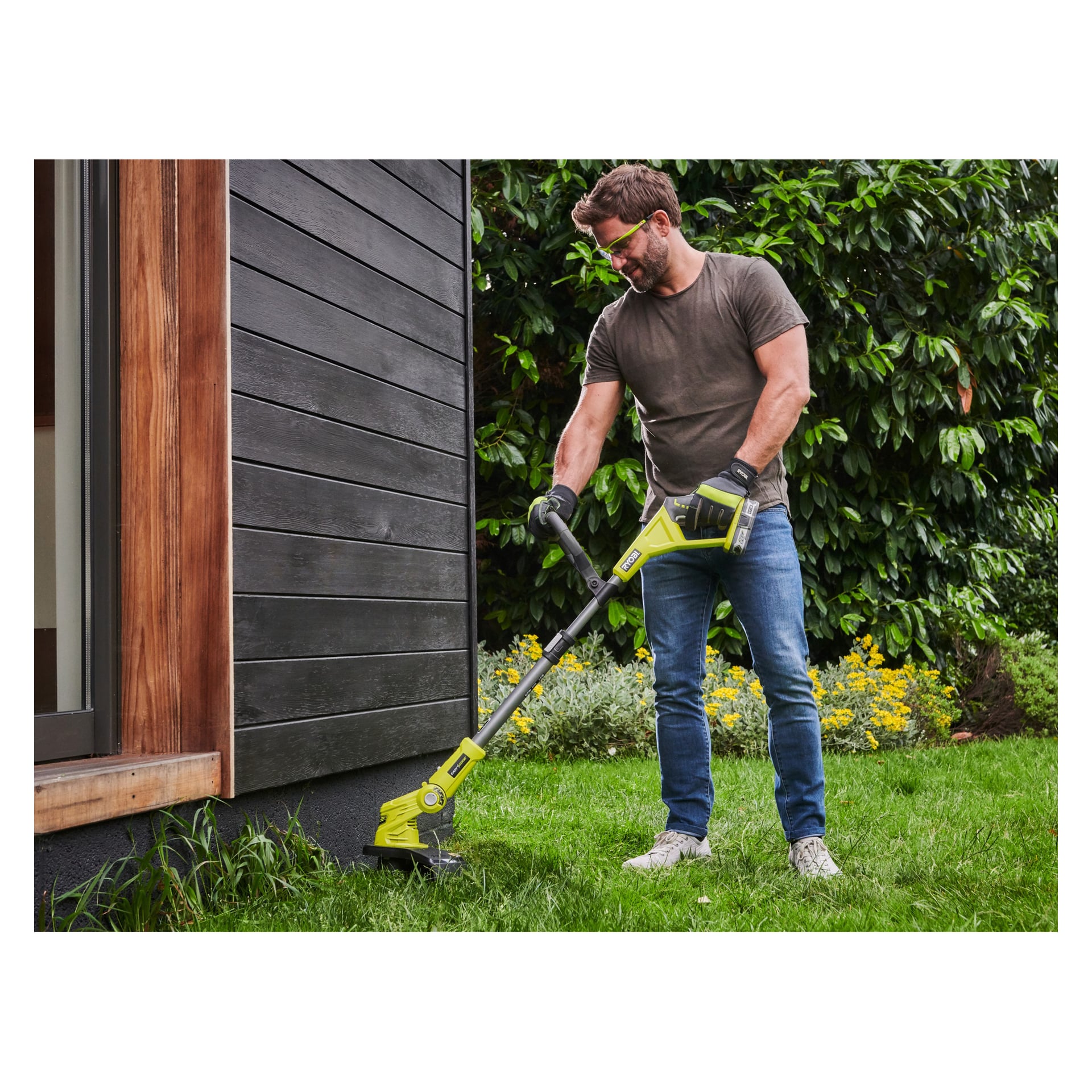En person som bär skyddsglasögon och handskar kör en gul Ryobi One+ batteridriven grästrimmer RLT183240F och klipper gräs längs kanten av ett hus med mörk träpanel. Scenen utspelar sig bland grönska och gula blommor.
