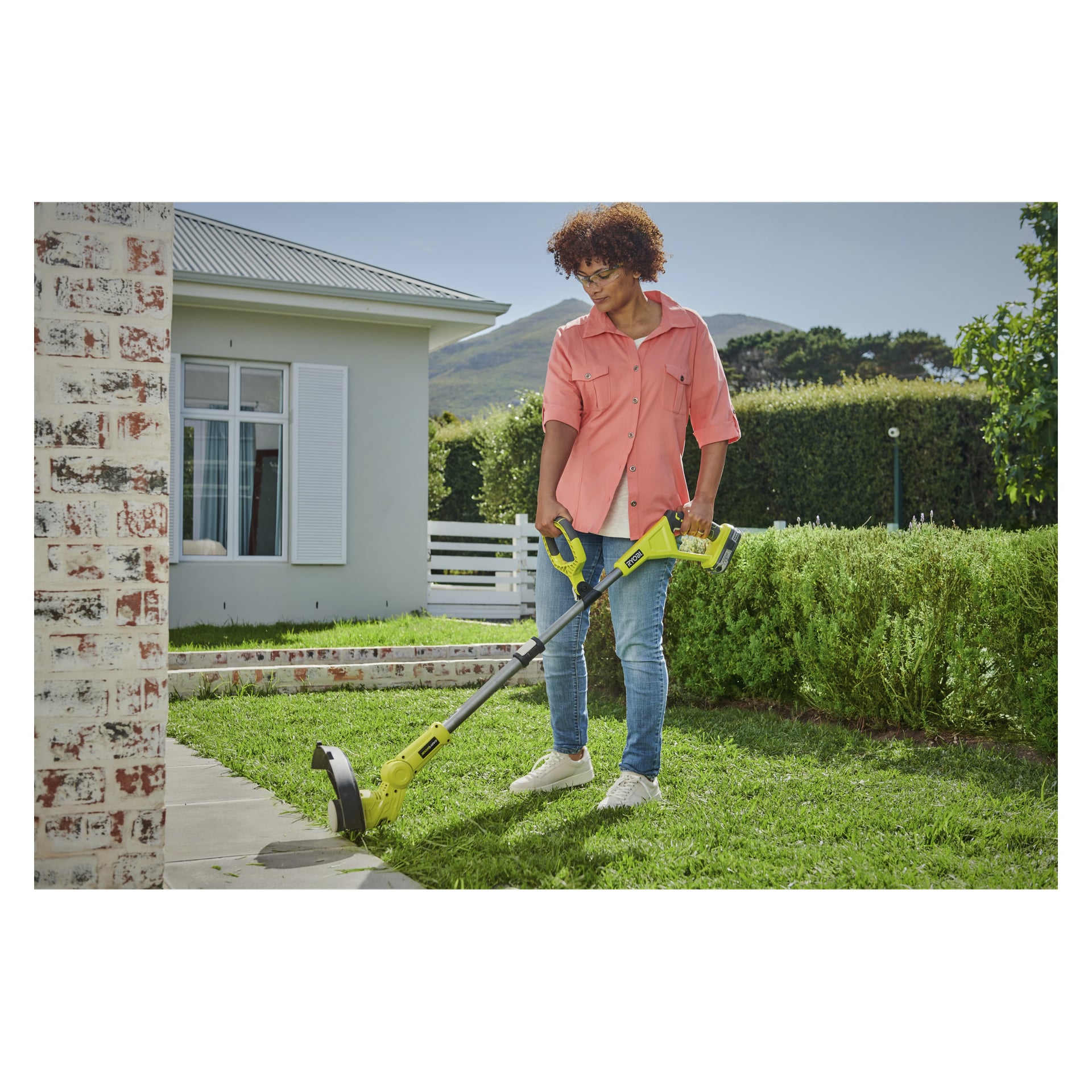 Någon i rosa skjorta, blå jeans och vita skor använder Ryobi One+ batteridriven grästrimmer RLT183240F för att trimma kanten av en gräsmatta bredvid en tegelvägg utomhus en solig dag.