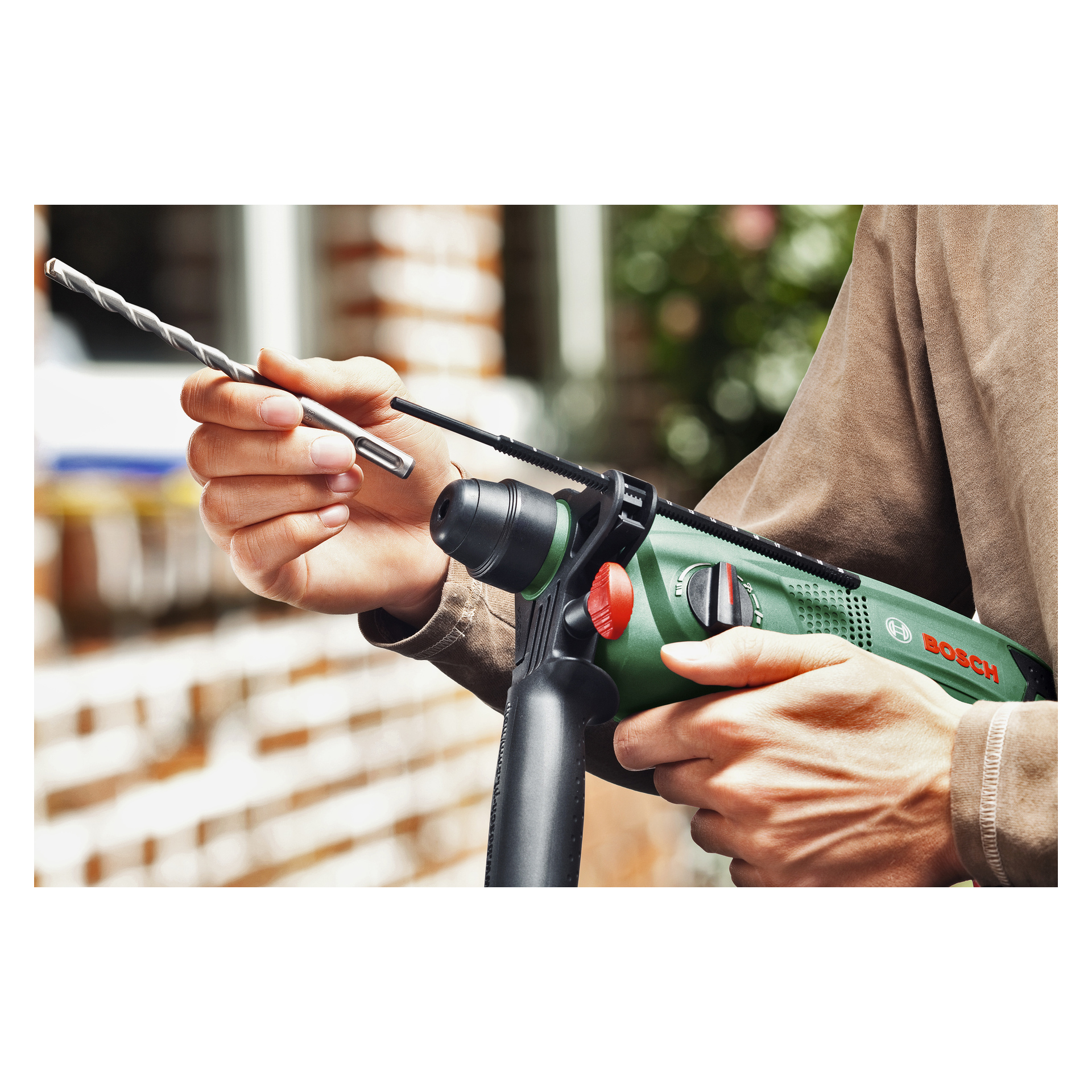 Joku pitää kädessään vihreää Bosch Hammer -porakonetta PBH 2100 RE, jossa on irrotettava sivukahva, valmistautuen asettamaan poranterän, kun tiiliseinä ilmestyy epätarkalle heidän takanaan.