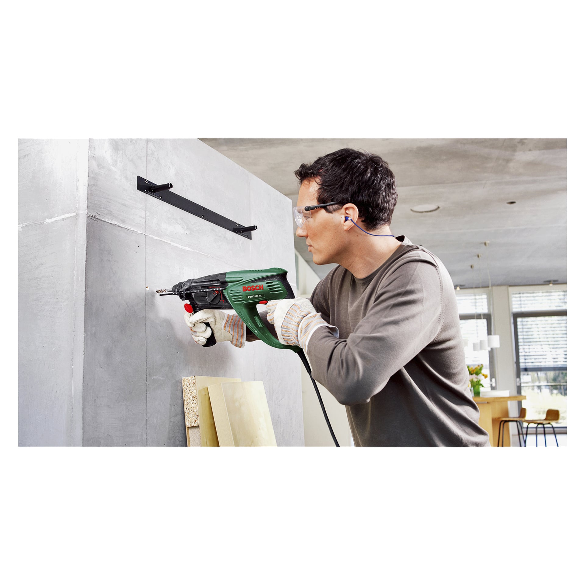 Mit Schutzbrille, Handschuhen und Gehörschutz bedient man den Bosch Hammer Drill PBH 2800 RE, um in eine Betonwand im Innenraum zu bohren. Mehrere Holzbretter stehen an der Wand in der Nähe.
