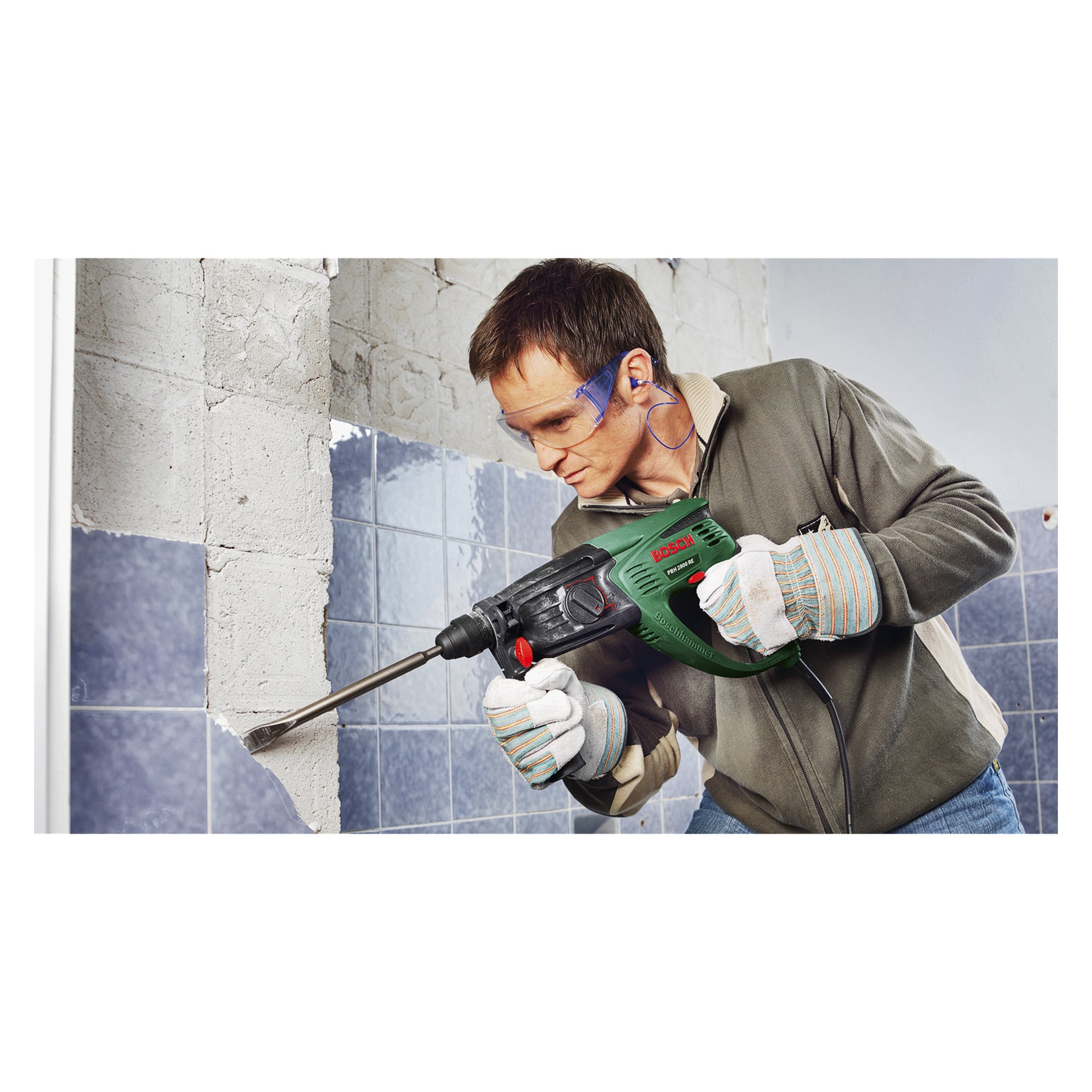 Mit Schutzbrille und Handschuhen bedient ein Mann einen grünen Bosch-Hammerbohrer PBH 2800 RE, um Ziegel von einer Wand abzuschlagen, die sich mitten in der Renovierung befindet, wodurch sowohl geflieste als auch kahle Bereiche freigelegt werden.