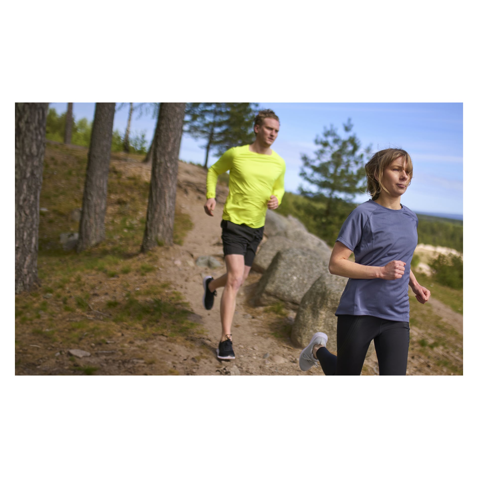Zwei Personen joggen auf einem Feldweg im Wald. Der Vorne trägt das graue Kayoba Women's Functional Top Tokyo mit schwarzen Leggings, während der Läufer dahinter ein gelbes Langarmshirt und schwarze Shorts trägt. Beide wirken konzentriert auf ihren Lauf.