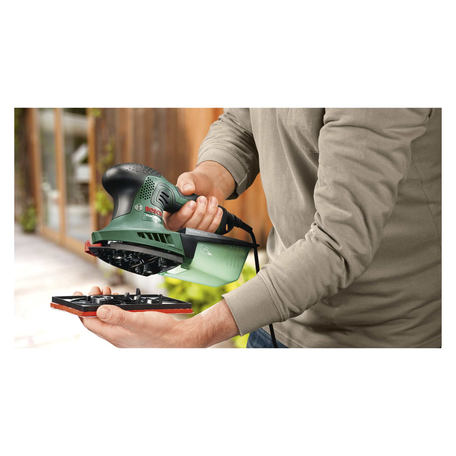Jemand hält mit einer Hand einen Bosch Multi-grind PSM 200 AES Schleifer und benutzt mit der anderen das Schleifpad. Der Hintergrund zeigt eine unscharfe Holzfläche, möglicherweise innen oder außen.