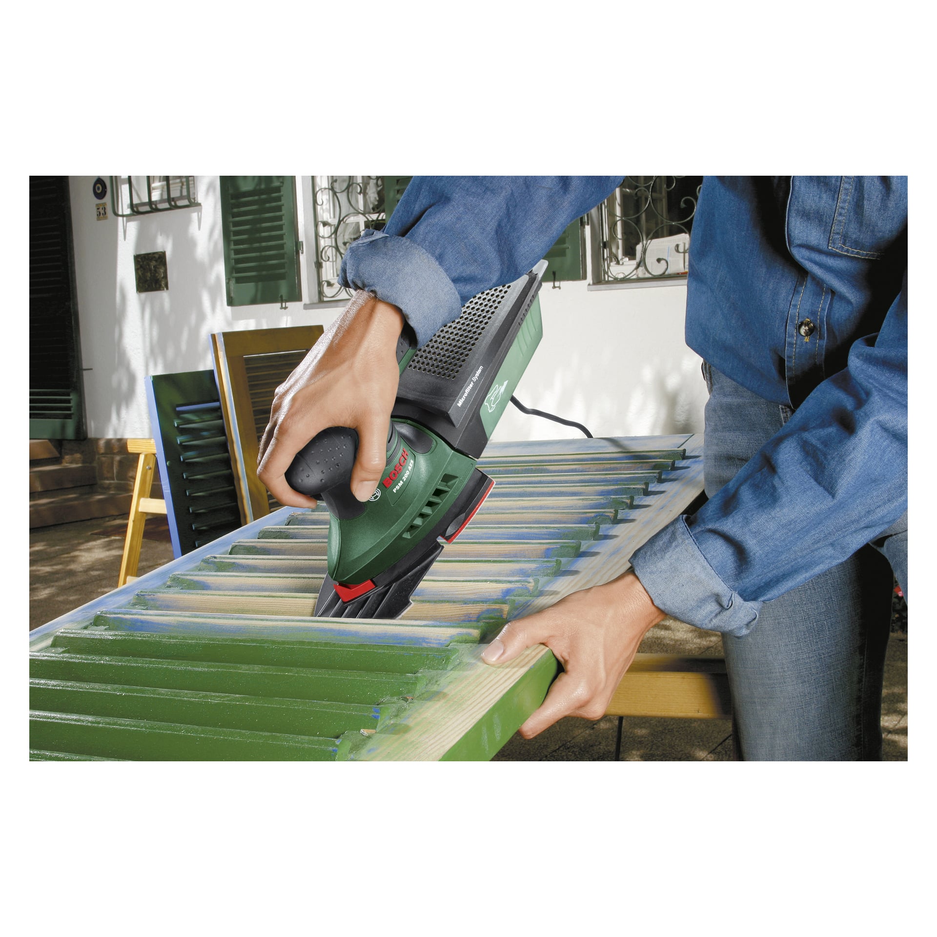 In einem blauen Langarmhemd bedient man einen Bosch Multi-grind PSM 200 AES Elektroschleifer, um grüne Farbe von einem Außenfenster zu entfernen. Während sie arbeiten, werden Stellen nacktem Holzes freigelegt, wo das Werkzeug effektiv die alte Farbe entfernt.