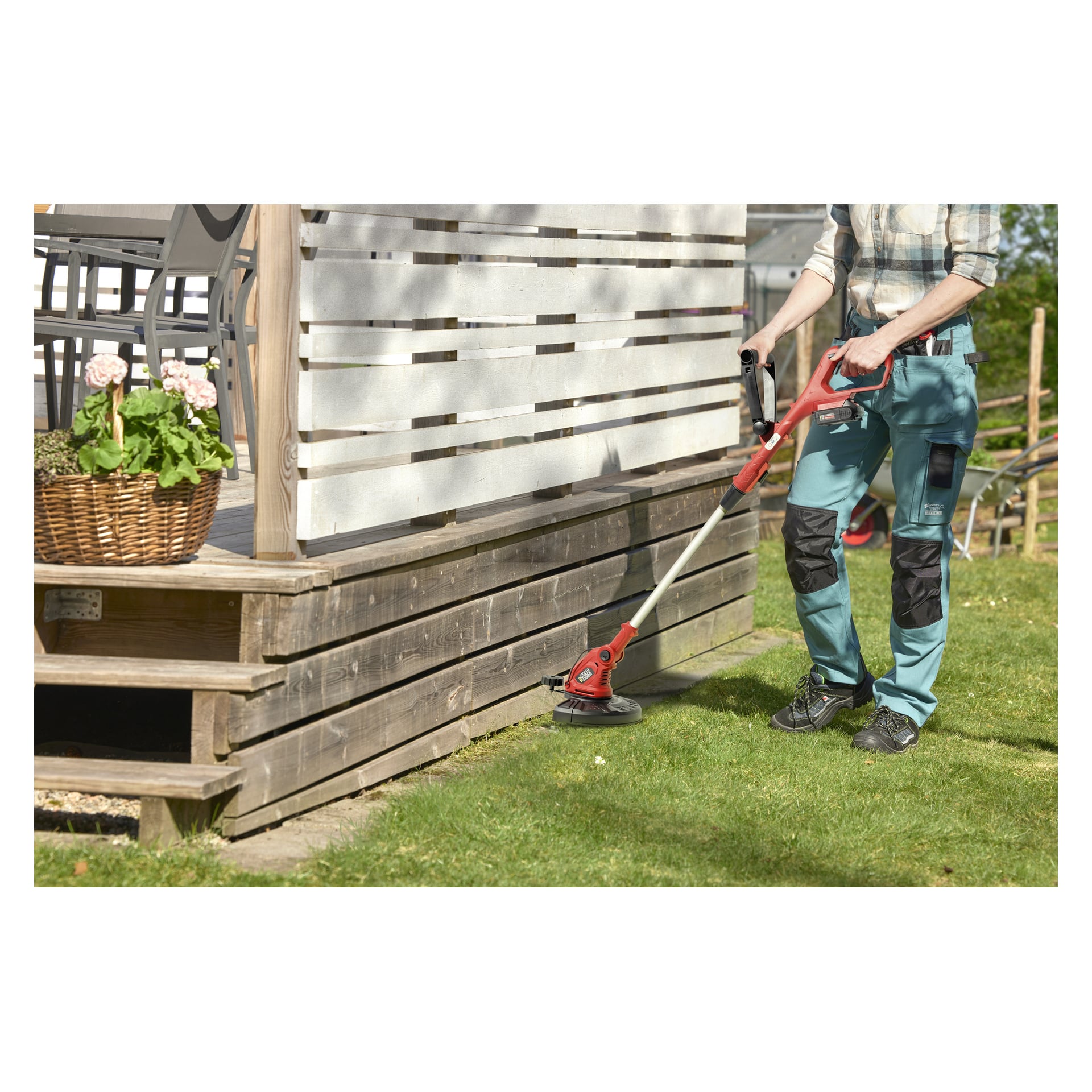 Någon klipper kanten på en gräsmatta bredvid en träaltan med vita paneler med hjälp av en Meec Tools Multiseries batteridriven grästrimmer GT-18V HOME 500. En korg av rotting med rosa blommor står på altanen, och det är en solig dag.