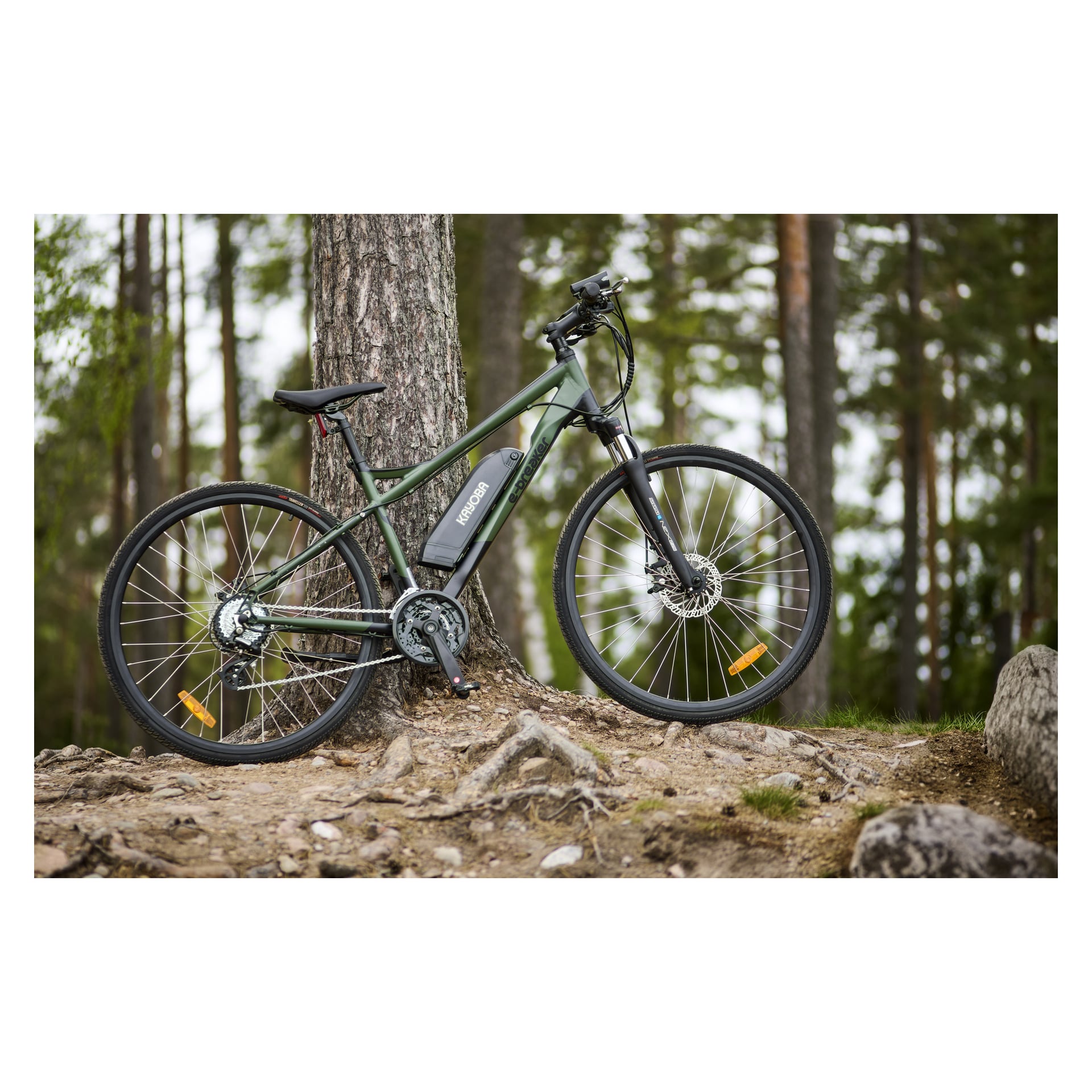 Ein Kayoba e-Breaker 2.0 Elektrofahrrad mit schwarz-grünem Farbschema steht an einem Baum in einem bewaldeten Bereich, umgeben von Steinen und Erde. Das Motorrad verfügt über robuste Offroad-Reifen und eine Batterie, die am Rahmen montiert ist.
