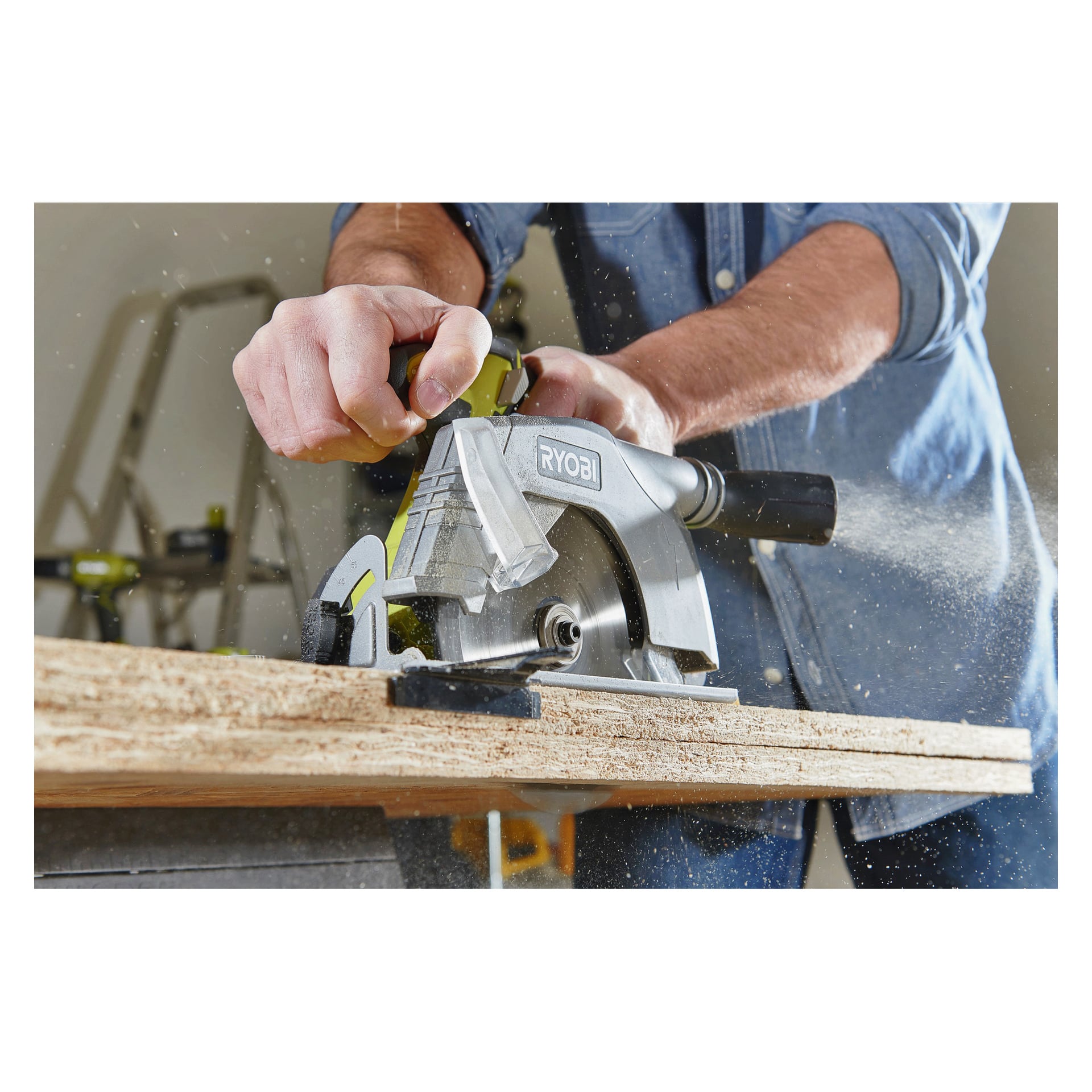 Mit einem Jeanshemd bedient jemand eine Ryobi One+ Kreissäge R18CS-140G, um durch ein Holzbrett zu schneiden, während Sägemehl verstreut wird. Eine Leiter und verschiedene Werkzeuge erscheinen im Hintergrund verschwommen.