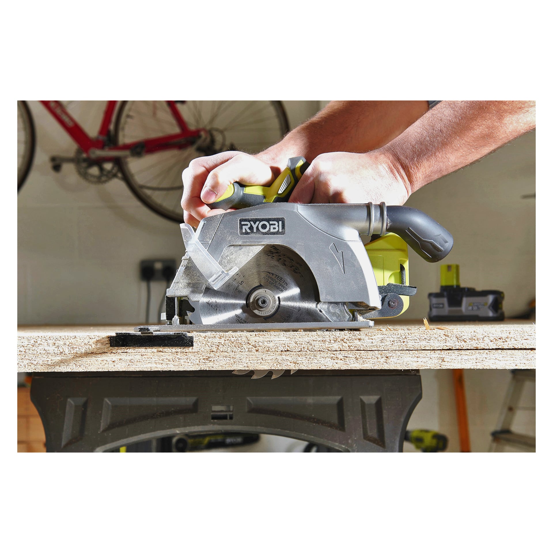 Man bedient eine Ryobi One+ Kreissäge R18CS-140G, um eine Holzplatte auf einer Werkbank zu schneiden. Im Hintergrund hängt ein rotes Fahrrad an der Wand. Die Person hält die Säge sicher mit beiden Händen.