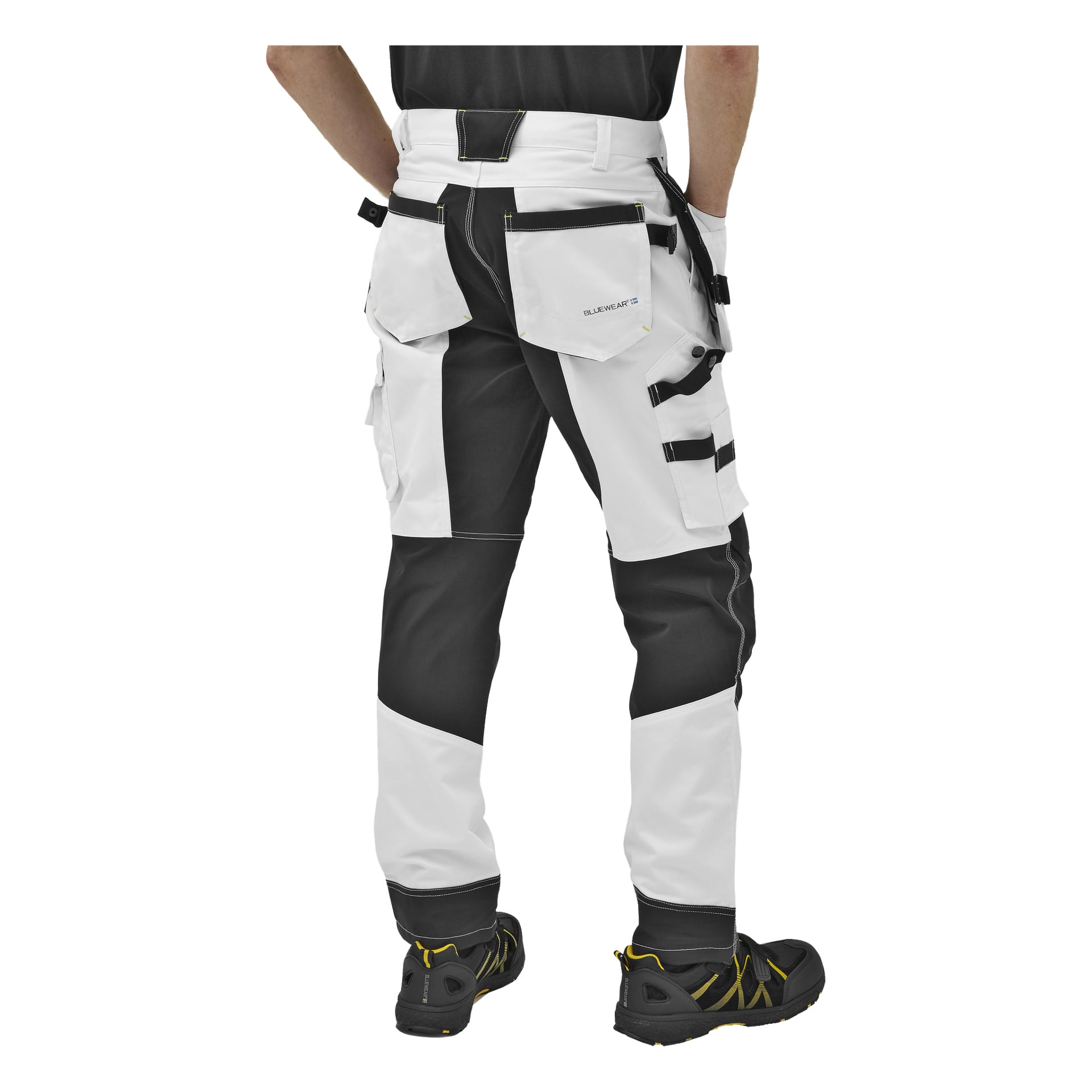 Patrząc od tyłu, osoba ubrana jest w męskie spodnie robocze Bluewear Men's Rex, w czarno-białych kolorach oraz z kilkoma kieszeniami. Noszą też czarne buty robocze z żółtymi akcentami. koszula tej osoby jest widoczna tylko częściowo.