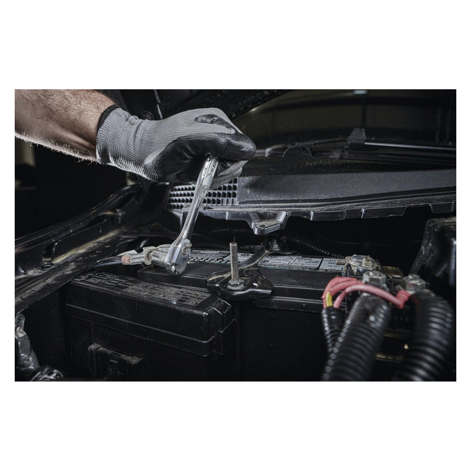 Eine behandschuhte Hand bedient einen DeWalt Box Spanner aus dem DWMT81610-0 50-teiligen Set, um eine Schraube an einem Batteriepol unter der Motorhaube zu lösen, umgeben von Batteriekabeln und Motorteilen.