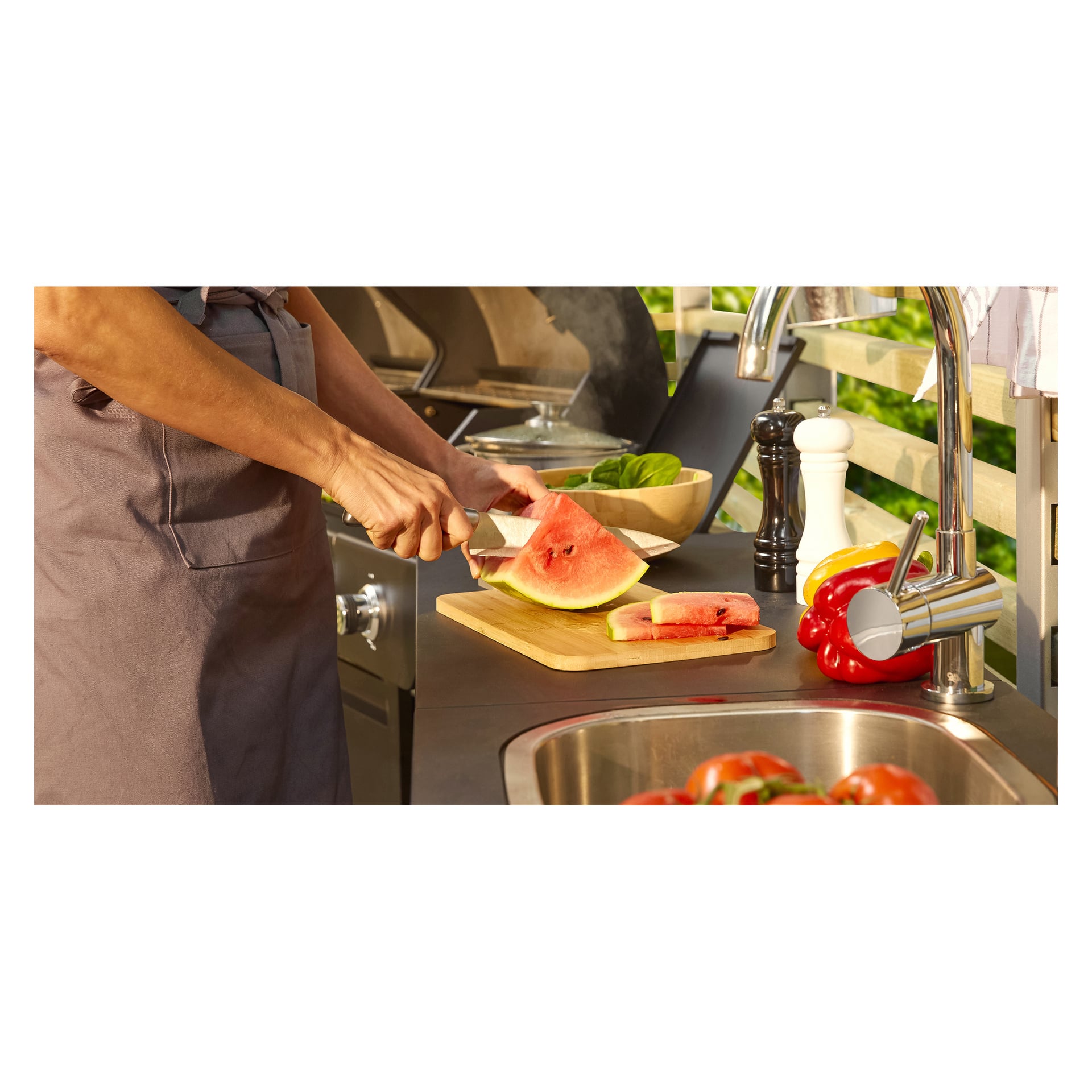Eine Person in einer Schürze schneidet eine Wassermelone mit einem Messer aus dem Menuett Knives + Messerblock 5-teilig, das auf einem hölzernen Schneidebrett in der Nähe des Waschbeckens steht, umgeben von verschiedenen Gemüsesorten sowie Salz- und Pfefferstreuern auf der Arbeitsplatte.