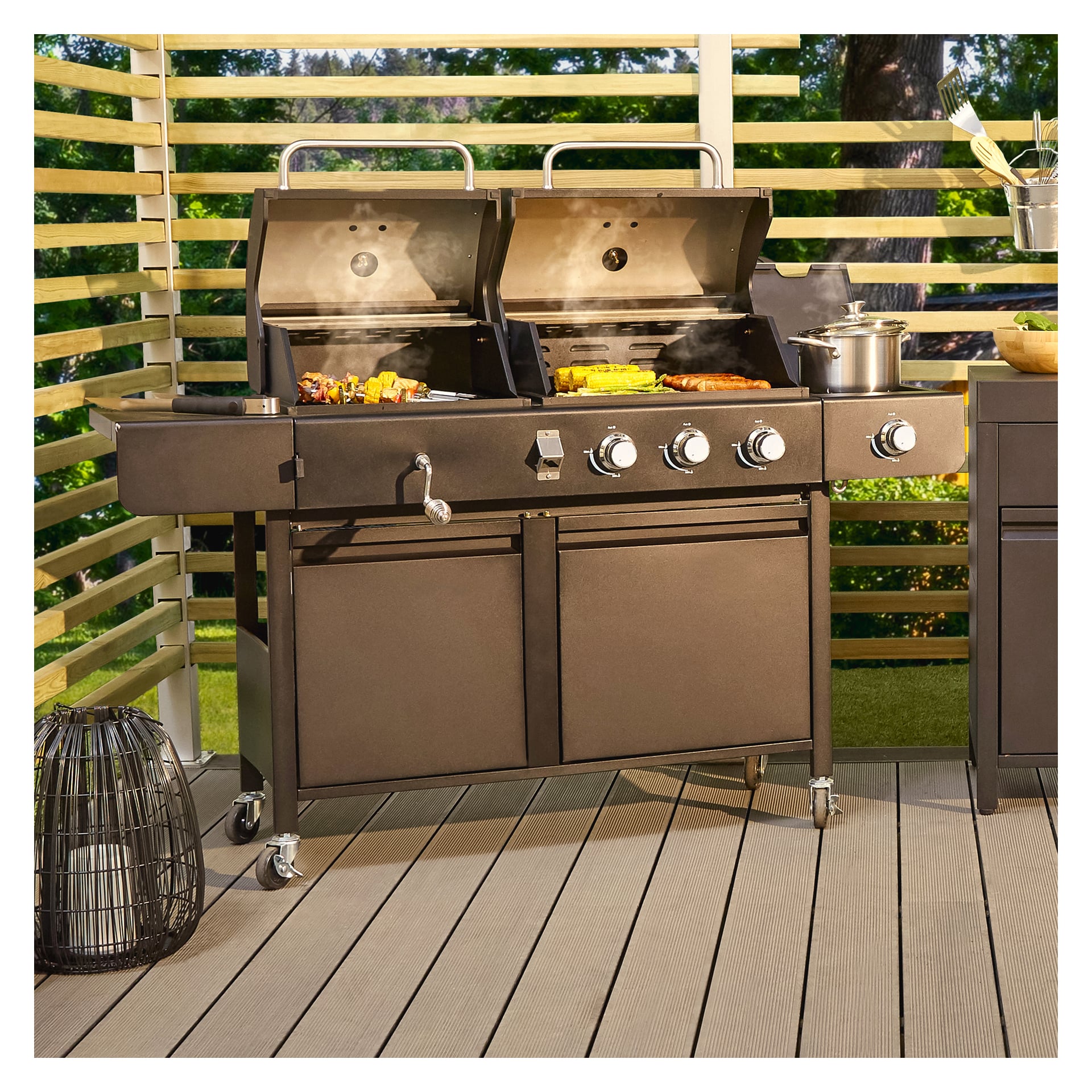 En Burns & Barkles All in One 8837 kol- och gasgrill med dubbla lock är placerad på en uteplats, båda grillytorna lagar mat med synlig ånga och en kastrull står i närheten. Framsidan har fem kontrollrattar, och grillen är omgiven av ribbel i trä och utemöbler.
