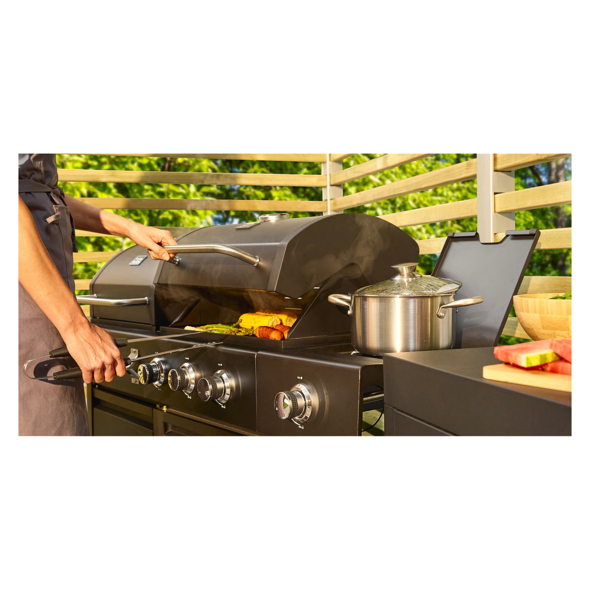 Någon öppnar locket på en Burns & Barkles kol-/gasgrill All in One 8837, vilket blottar olika grönsaker och majs inuti. På grillens sidobrännare står en kastrull i rostfritt stål. Uppställningen är placerad på en trädäck omgiven av frodig grönska.
