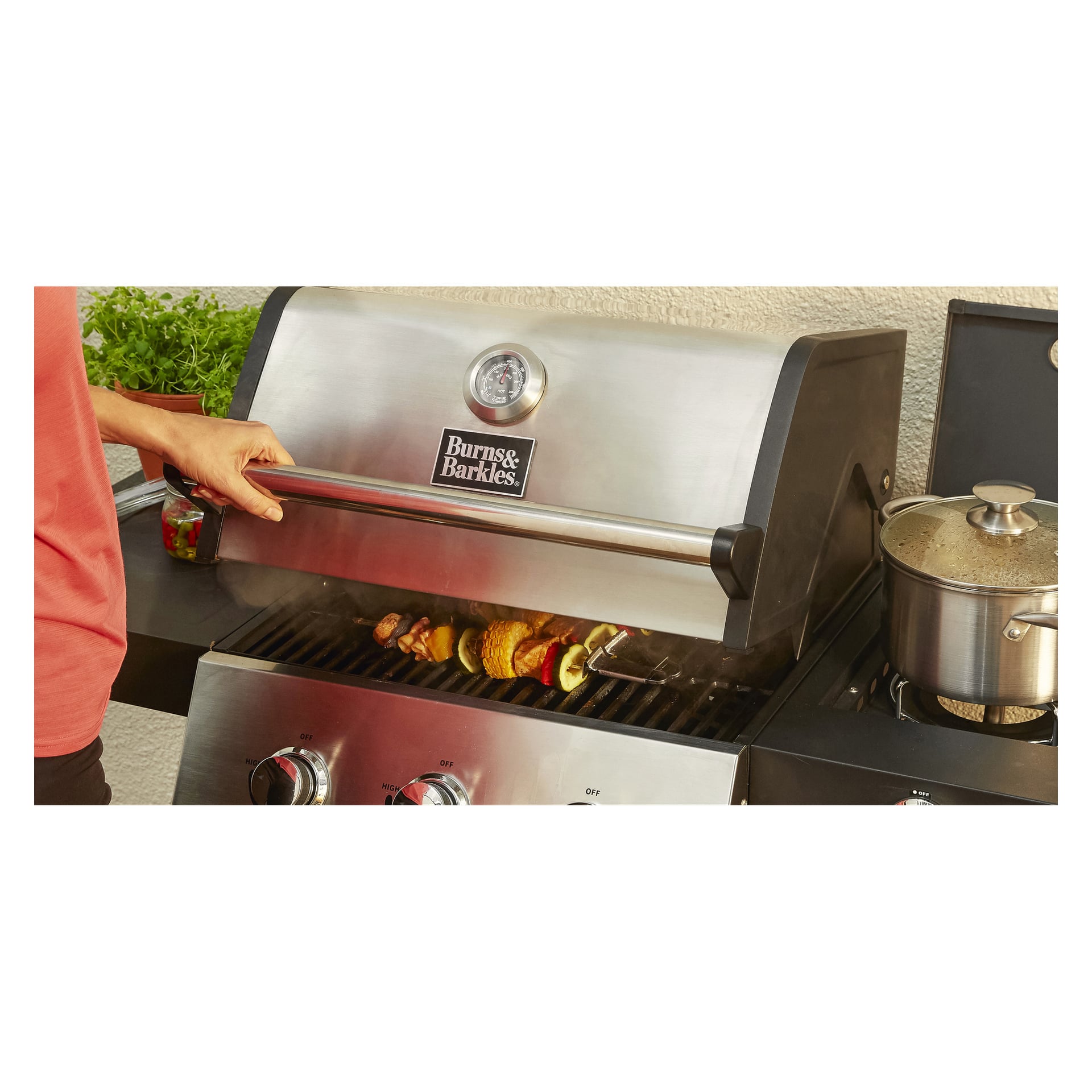 Någon lyfter på locket på en Burns & Barkles Gas Grill Steely King 5042B, vilket avslöjar spett med grönsaker och köttgrillning inuti. En kastrull står på den intilliggande sidoplattan.