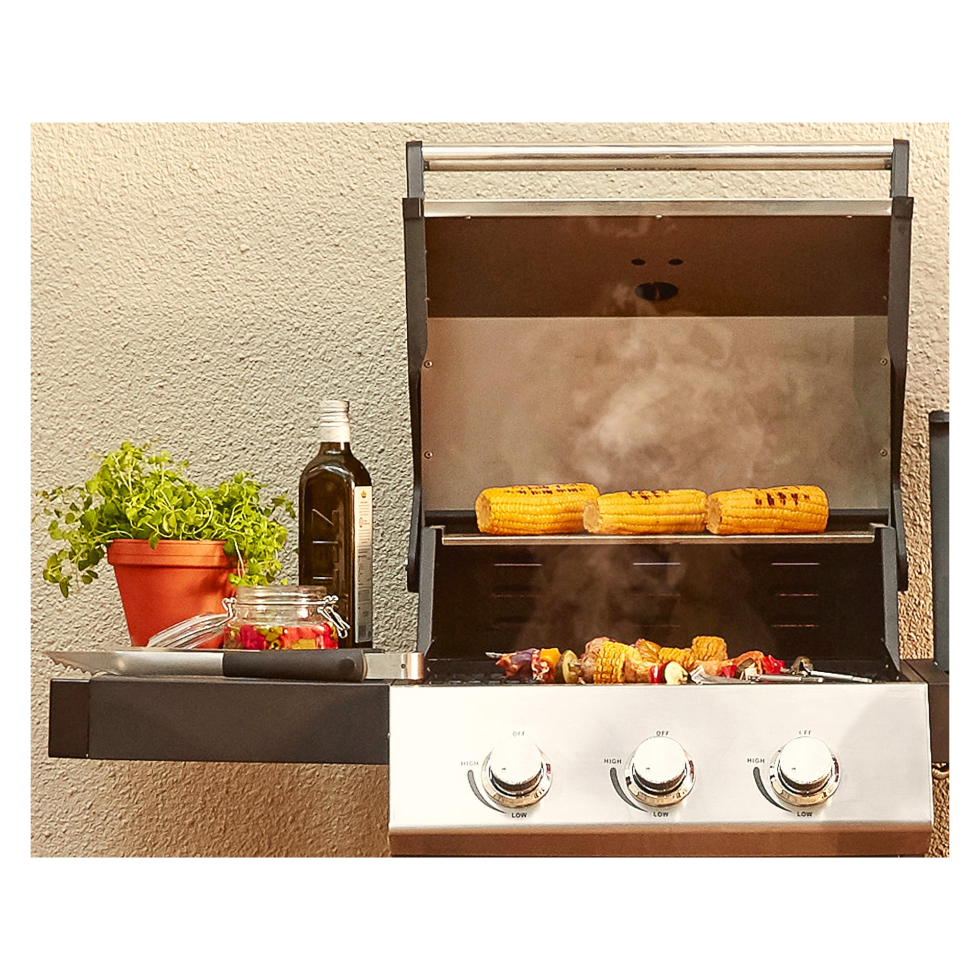Maiskolbe freser på en gassgrill med damp som stiger opp, og spyd med fargerike grønnsaker grilles under. På det tilstøtende sidebordet er gjenstander som en pottet urt, en kniv, en flaske olje og en Menuett-glasskrukke med gyngestopp ordnet i orden.
