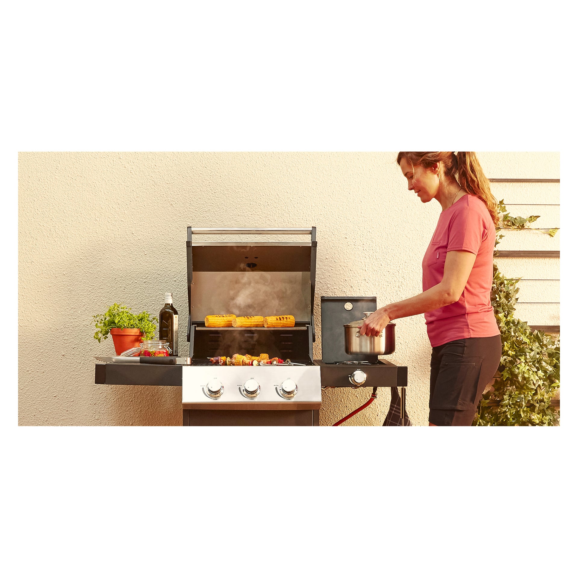 En kvinna i rosa skjorta grillar grönsaker och majs på Burns & Barkles Steely King 5042B gasgrill. Hon håller en kastrull över sidobrännaren, med färska örter och en flaska olja placerad på grillens sidohylla bredvid en beige vägg.