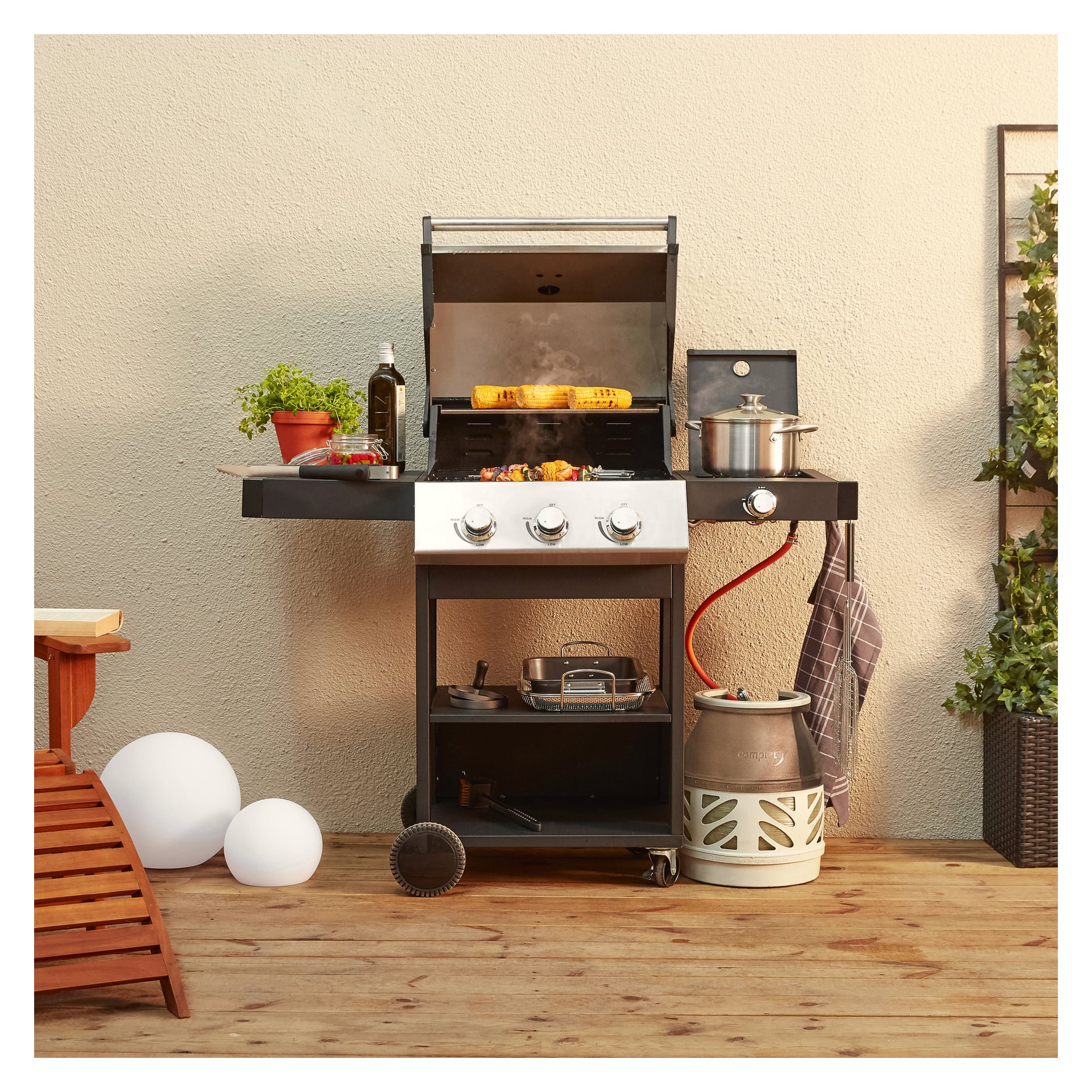 En Burns & Barkles Gas Grill Steely King 5042B med tre plattor, en kastrull, brödbitar på grillen, färska örter, olivolja och olika redskap står på en träbalkong intill en vägg. I närheten finns en propantank, en bänk, en krukväxt och två vita sfäriska föremål.