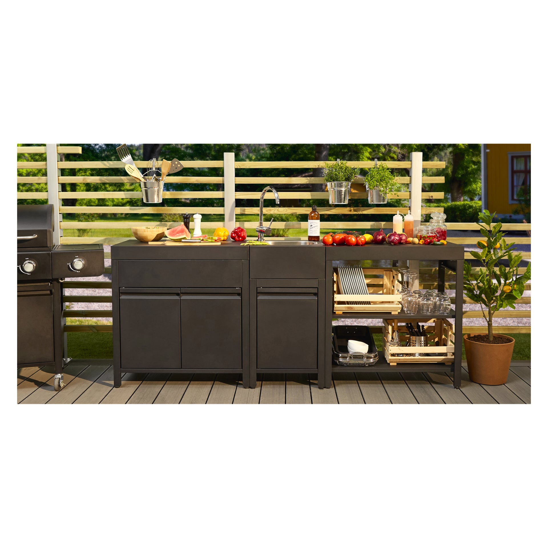 Warsztat Burns & Barkles z szafką Outdoor Kitchen 8044B jest wyróżniony, wyposażony w nowoczesną czarną jednostkę zlewową. Półki są starannie uporządkowane z różnymi przyborami kuchennymi, świeżymi warzywami i owocami, butelką wina oraz naczyniami. Po prawej stronie stołu warsztatowego stoi doniczkowa roślina. Cała scena rozgrywa się przed drzewami i jest otoczona drewnianym ogrodzeniem z listew.