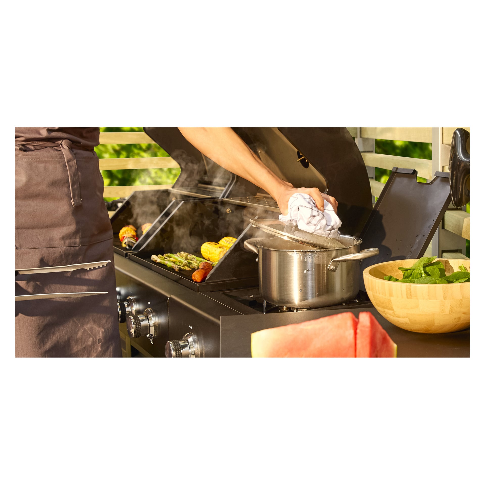 Henkilö esiliinassa grillaa vihanneksia ja maissia, pitäen toisessa kädessään grillaustyökalua ja toisella nostamassa höyryävän Menuett-kattilan kannen. Läheisellä tasolla on kulhollinen pinaattia ja viipale vesimelonia.