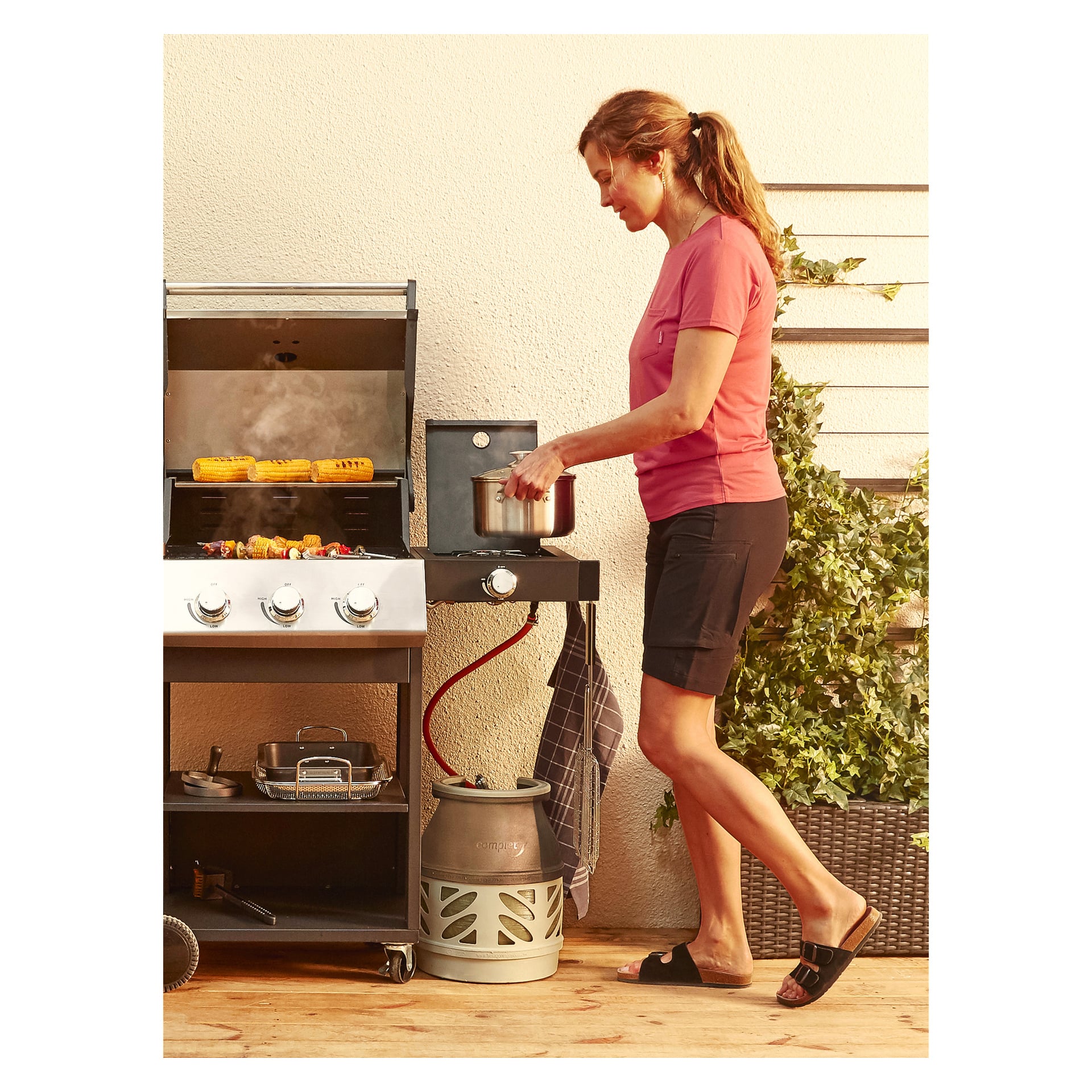 En kvinne iført rosa skjorte og svarte shorts står ute ved siden av en gassgrill og bruker Burns & Barkles Grill Pan på en sideplate. Grønnsakene koker på grillen, og en propantank er synlig under mens hun lager mat nær potteplanter.