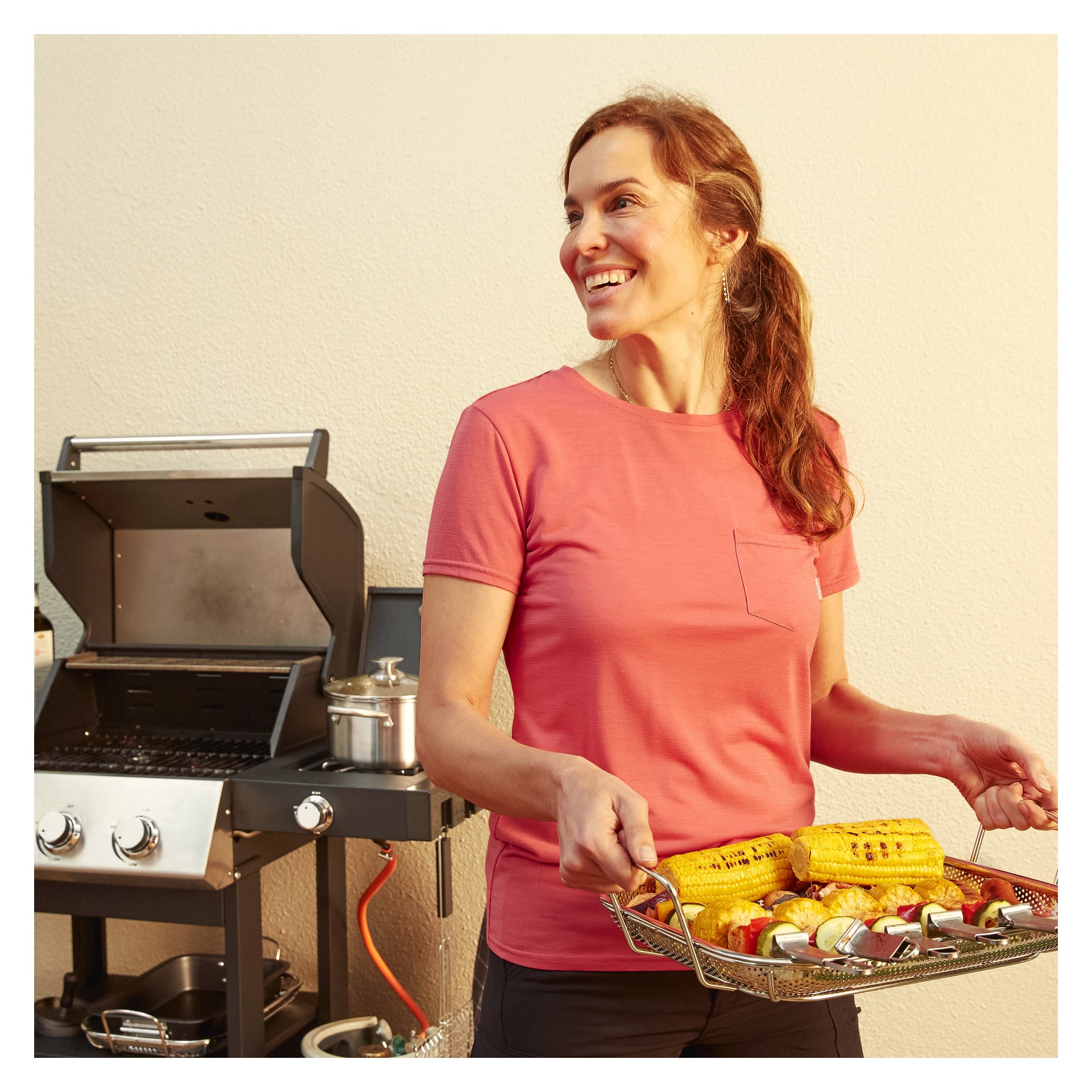 En smilende kvinne i rosa skjorte står ved siden av en grill med en gryte på toppen, holder Burns & Barkles Grill Pan fylt med mais og spyd mens hun ser til siden.
