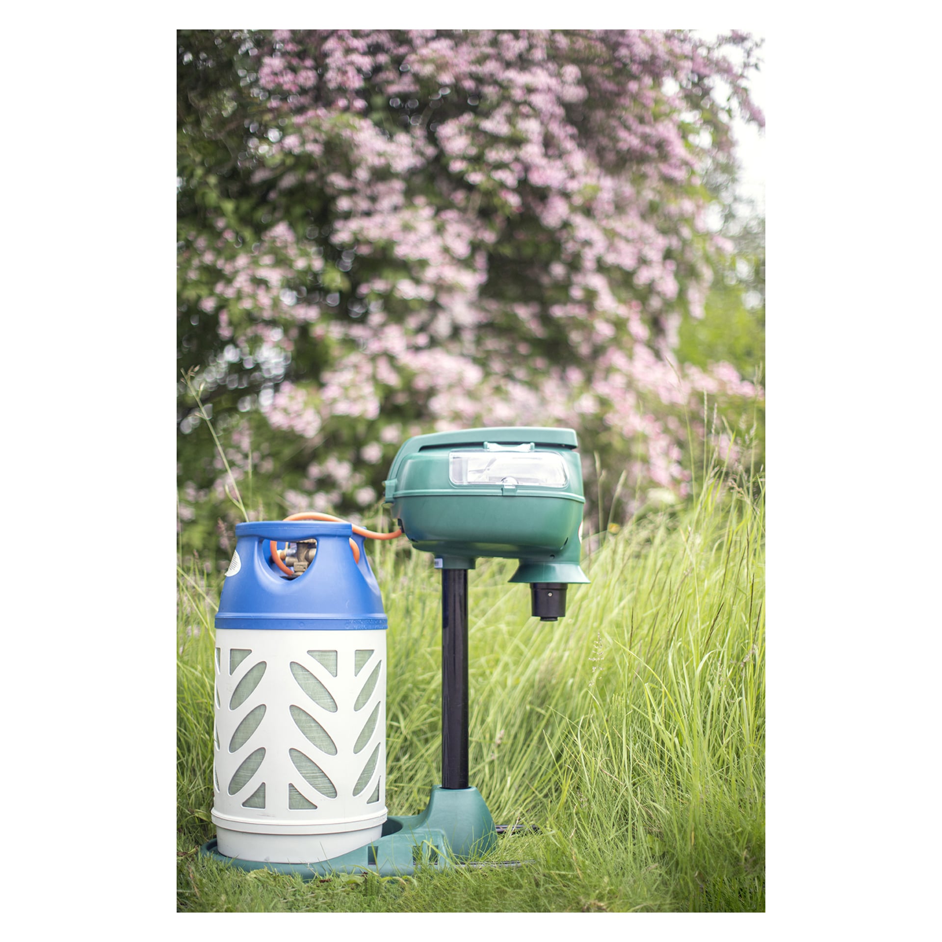 En blå och vit propantank, kopplad med en slang, driver Mosquito Magnet Pioneer – en grön myggfälla från Mosquito Magnet – placerad på en svart bas omgiven av högt gräs och blommande buskar i bakgrunden.