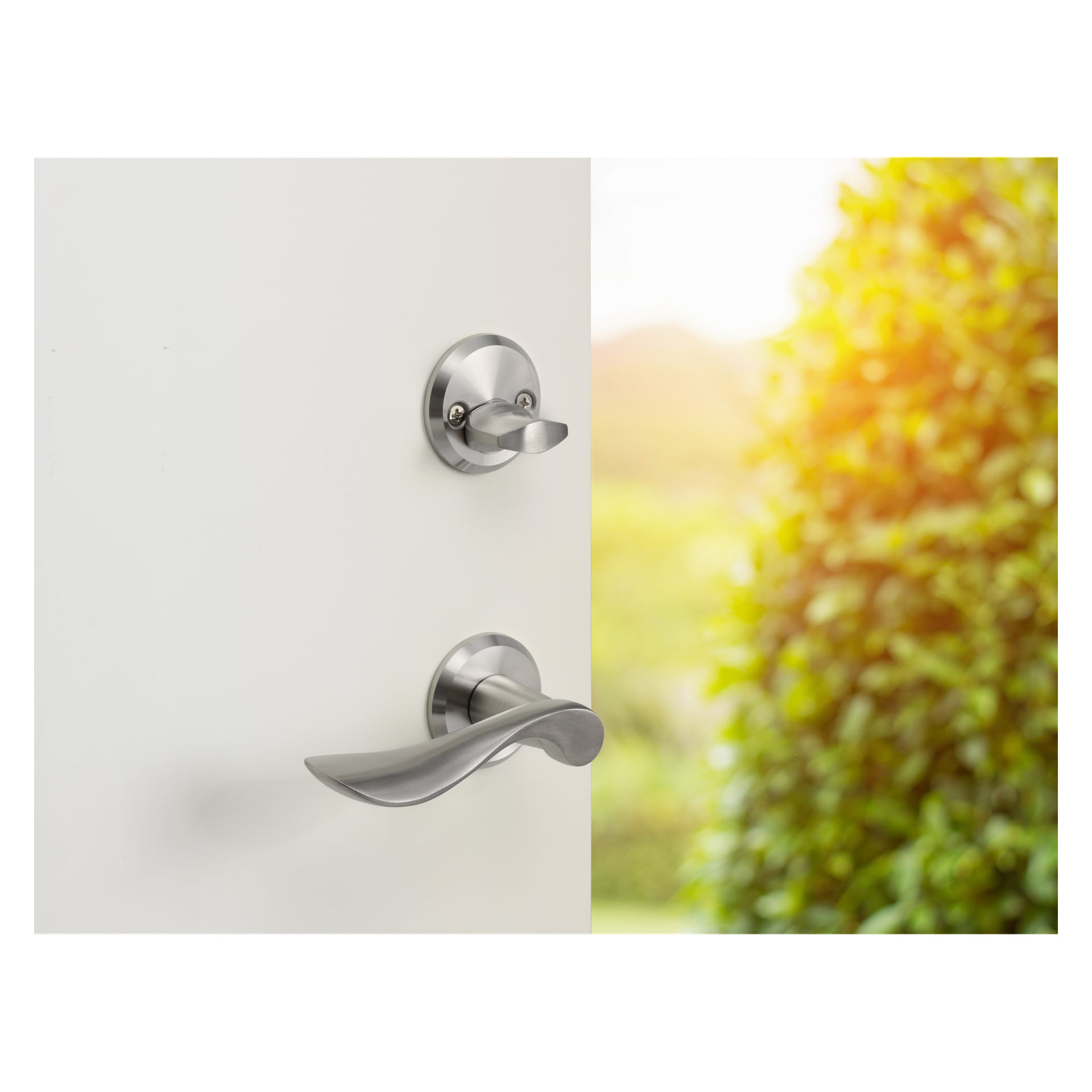 En hvit dør med en børstet nikkel Hard Head Front Door Handle står på gløtt, og viser en solfylt utsikt over grønt løvverk utenfor.