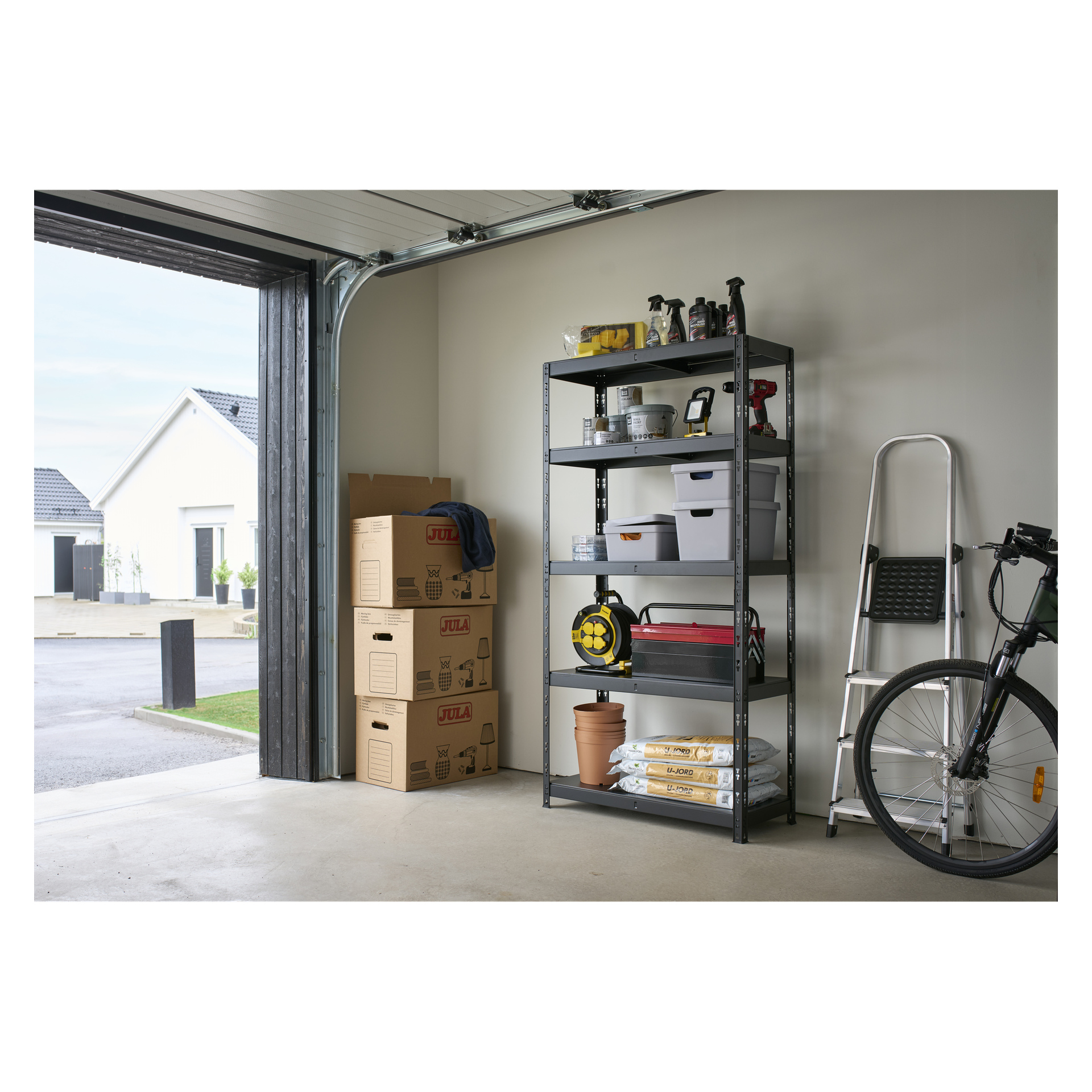 Inne i ett garage med öppen dörr finns en Hard Head Storage-hylla med ett urval verktyg, lådor och trädgårdsartiklar. I närheten finns en cykel, en hopfälld stege och flera flyttlådor staplade tillsammans. Genom den öppna ingången kan hus från bostadsområdet ses utomhus.