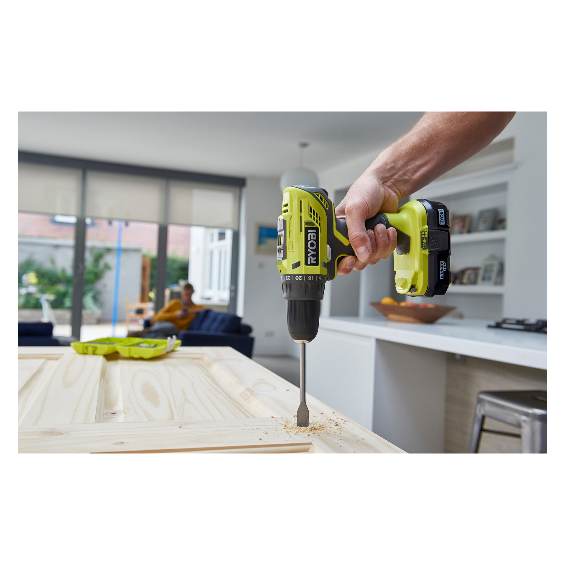 Yksityiskohtainen kuva henkilöstä, joka käyttää Ryobi One+ -ruuvimeisseliä R18DD32-0 -ruuvimeisseliä poratessaan puupalaa tyylikkäässä keittiöympäristössä, kun taas toinen henkilö istuu sinisellä sohvalla heidän takanaan.