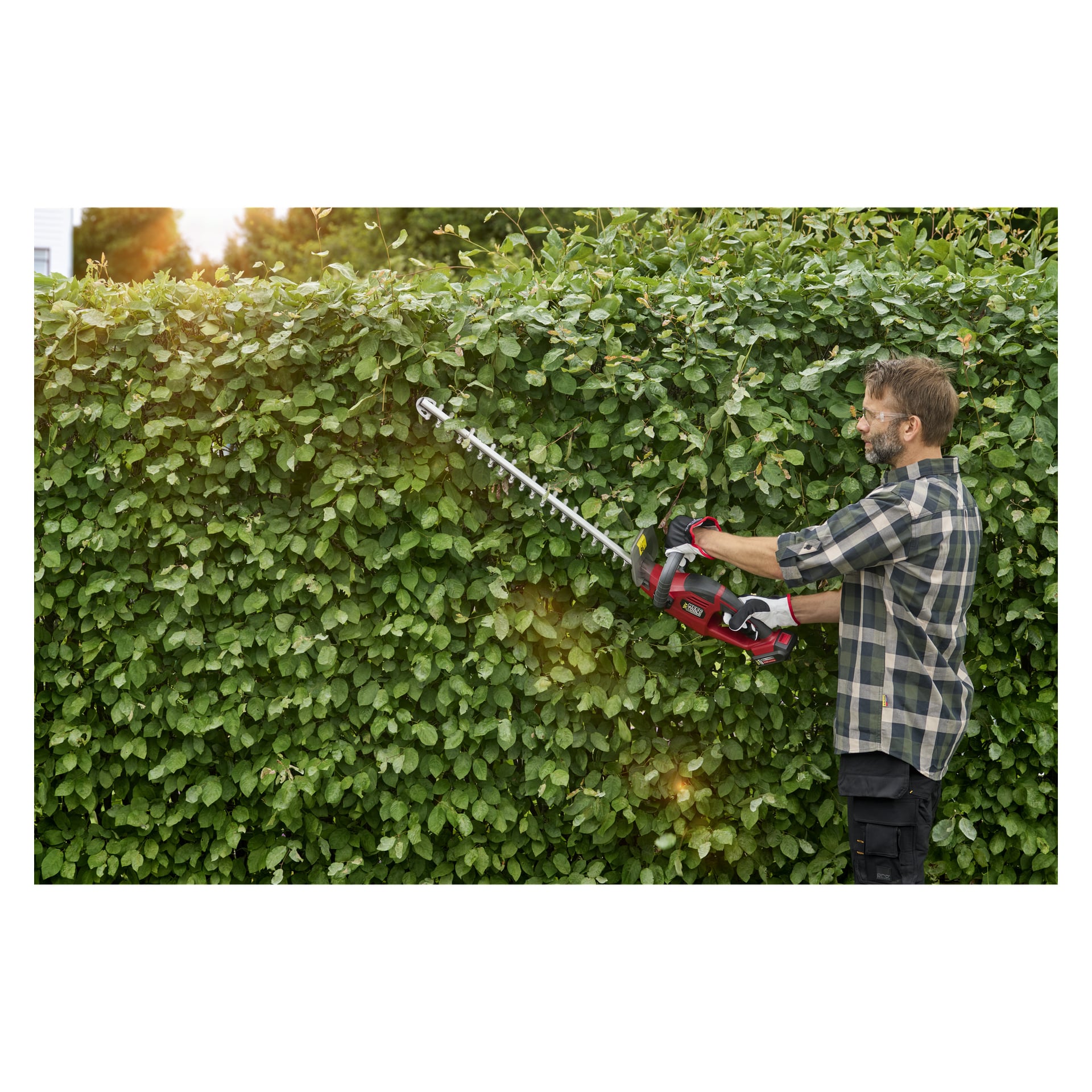 En mann med vernebriller, hansker og rutete skjorte pynter en høy grønn hekk utendørs med Meec Tools Multiseries batteridrevne hekktrimmer HT-18V HOME 500 i sterkt sollys.