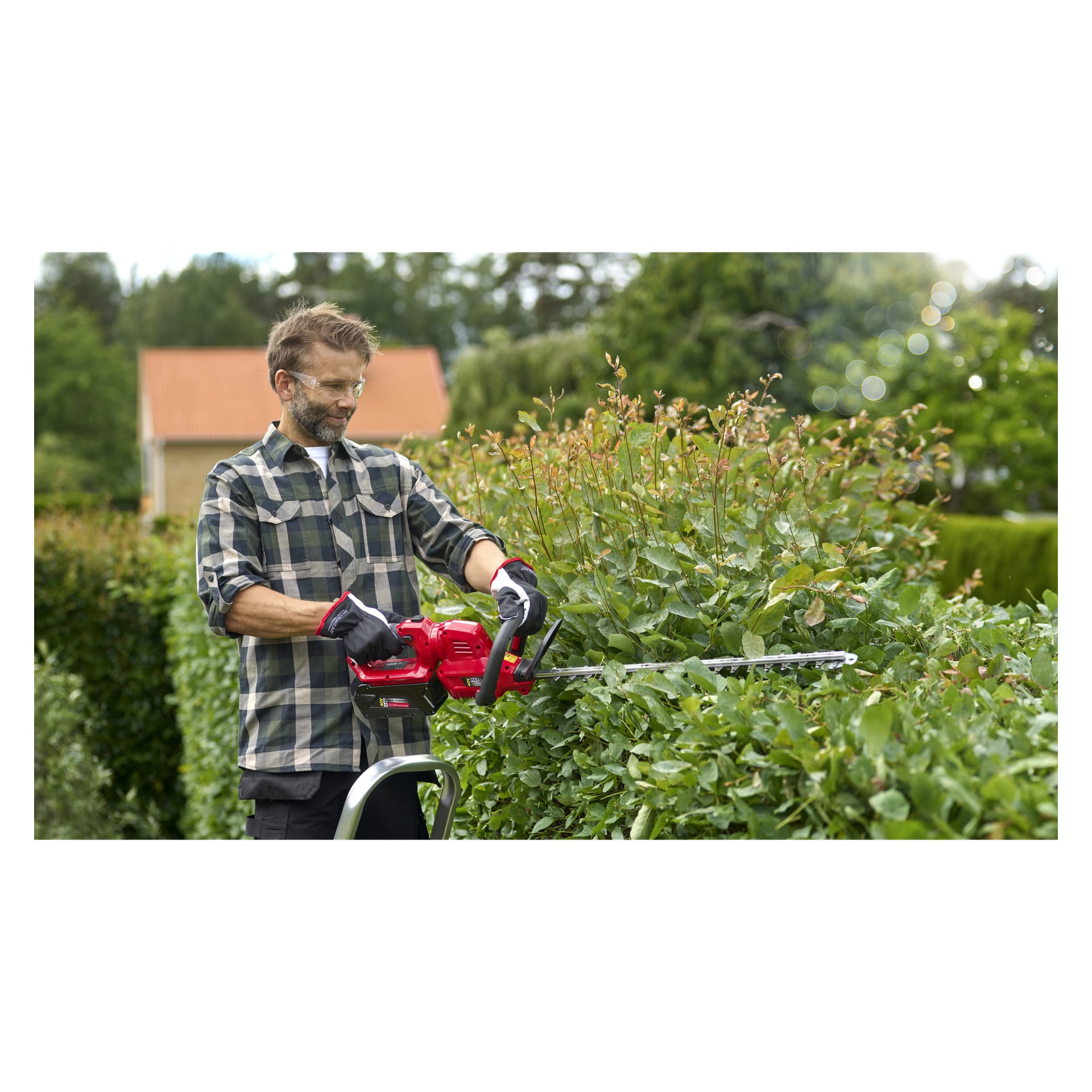 Mężczyzna w koszuli w kratę, okularach ochronnych i rękawiczkach używa podtrzymywacza Meec Tools Multiseries 40 V zasilanego bateriami, aby uformować bujny zielony żywopłot w ogrodzie. Za nim stoi dom i kilka drzew.