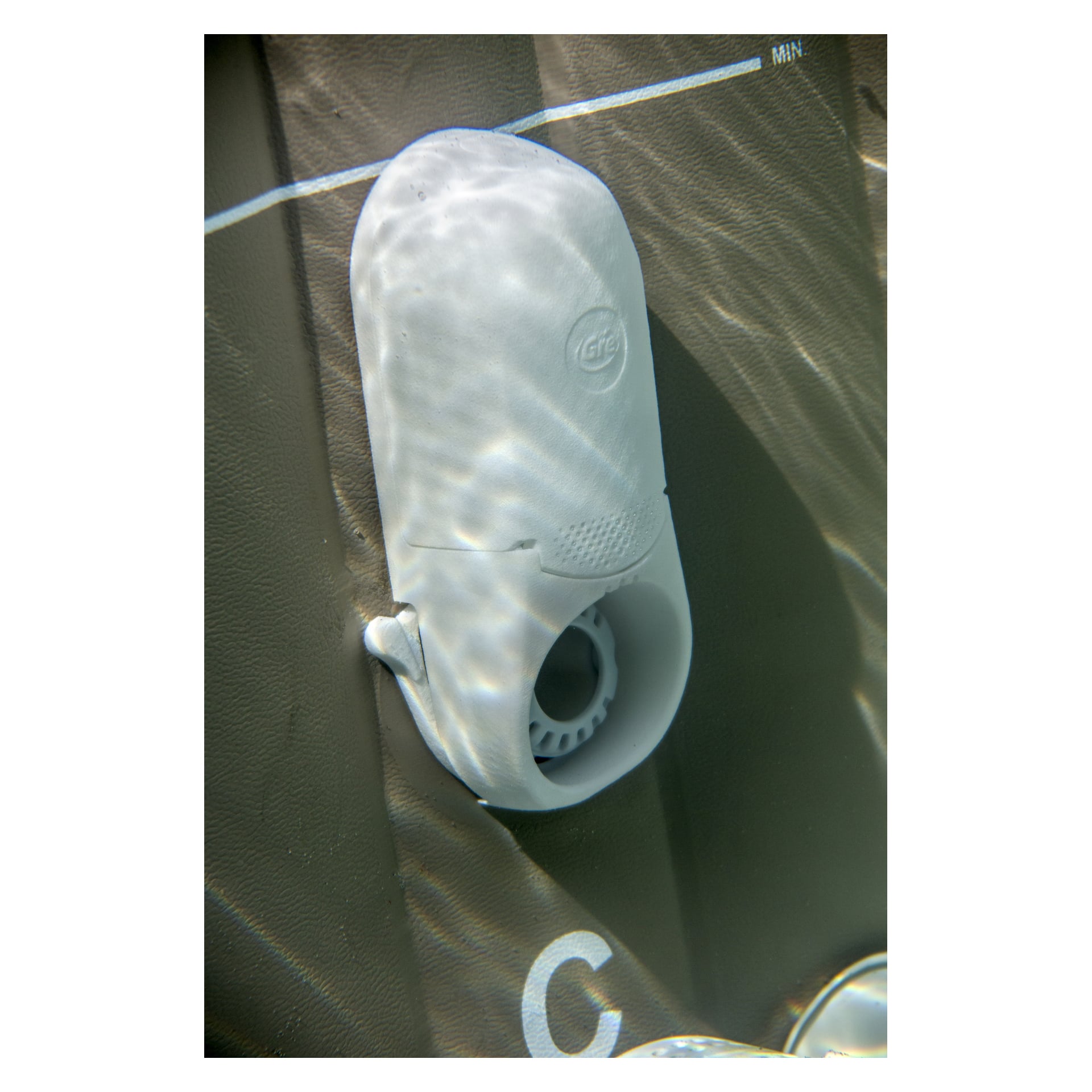 Ett vitt plastkit för SPA CAPS Spa Bath Disinfection Kit från Swim Fun är monterat på poolväggen och delvis nedsänkt. Mätmarkeringar är synliga på väggen, med skimrande ljusmönster från vattnet som reflekteras på ytorna.