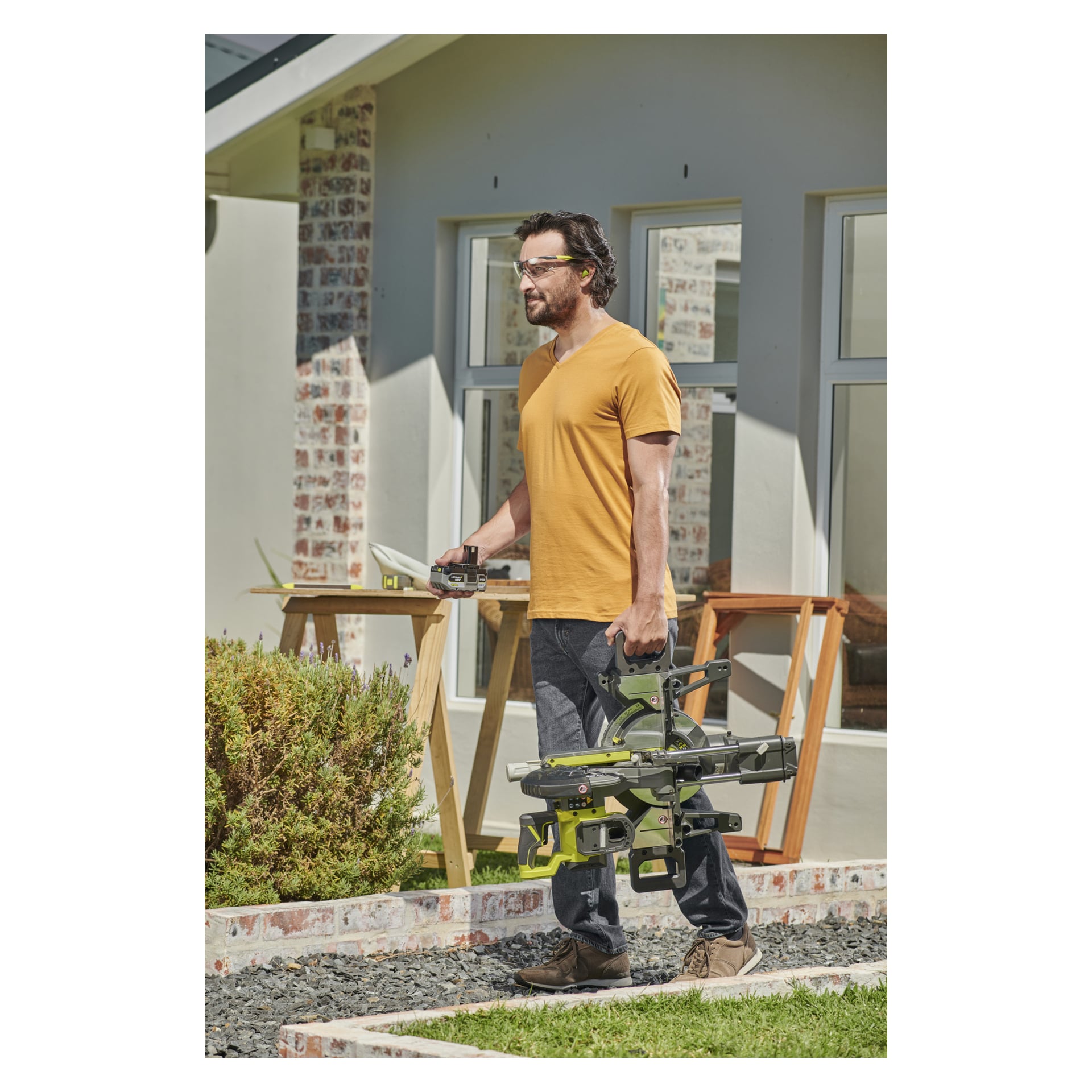 En man med skyddsglasögon och orange T-shirt bär en Ryobi One+ batteridriven mitresåg RMS18190-0 tillsammans med ett måttband utomhus bredvid ett hus, med träsågar och ramar synliga bakom sig.