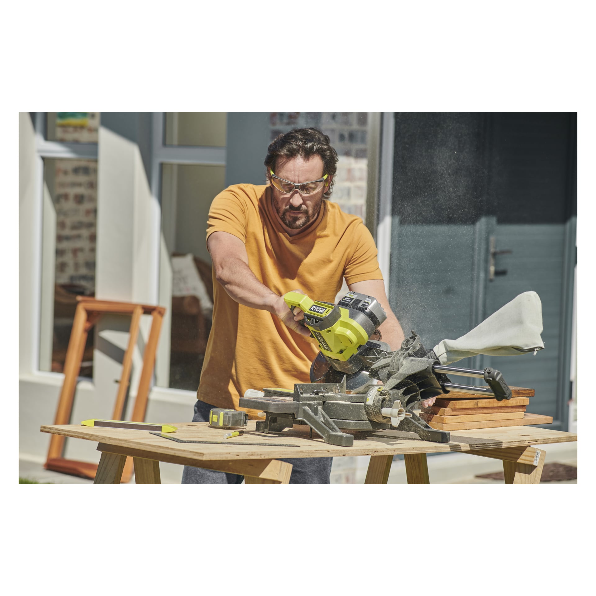 En man med skyddsglasögon och orange t-shirt använder en batteridriven Ryobi One+ geringssåg (modell RMS18190-0), där han sågar trä på ett utebord. Bordet innehåller ytterligare byggverktyg och träplankor, med ett hus i bakgrunden.