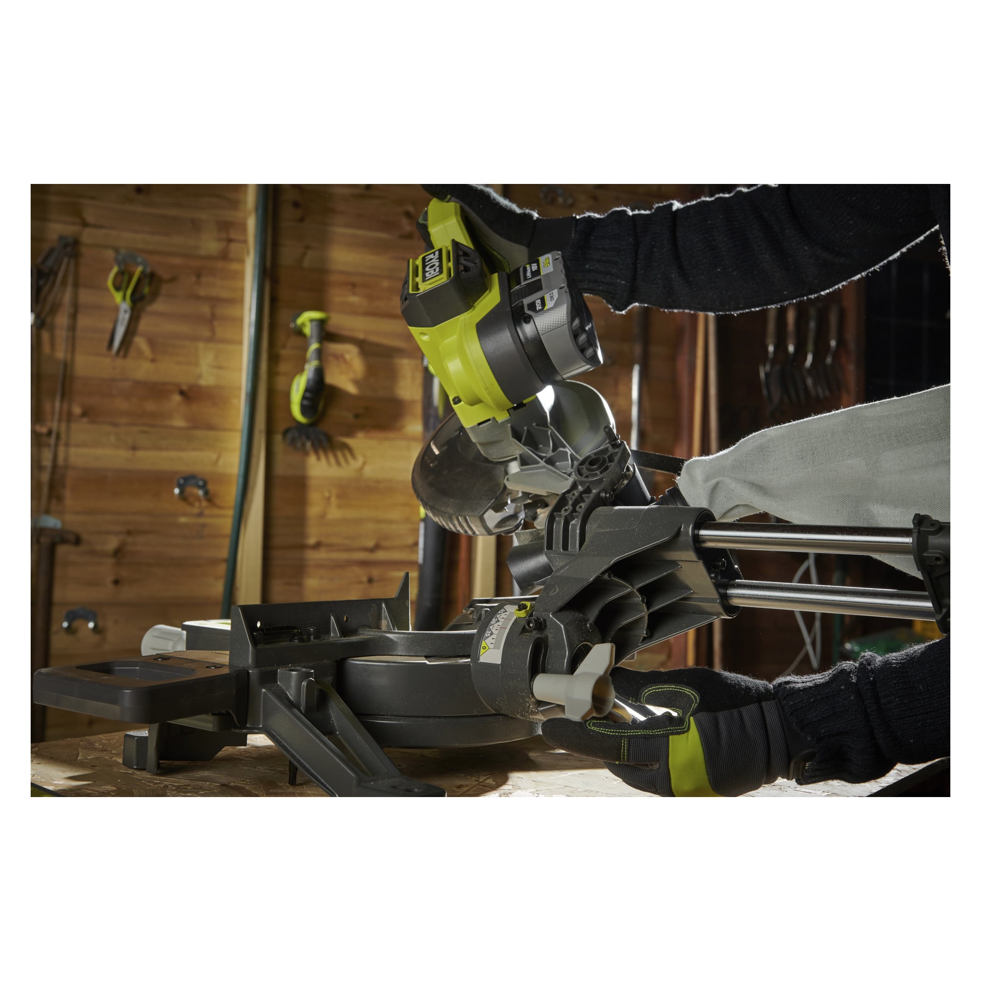 Handskbeklädda händer i mörka ärmar styr en Ryobi One+ batteri-geringssåg RMS18190-0 för att såga en träbit i en verkstad i trä. Olika verktyg är organiserade på väggen bakom.