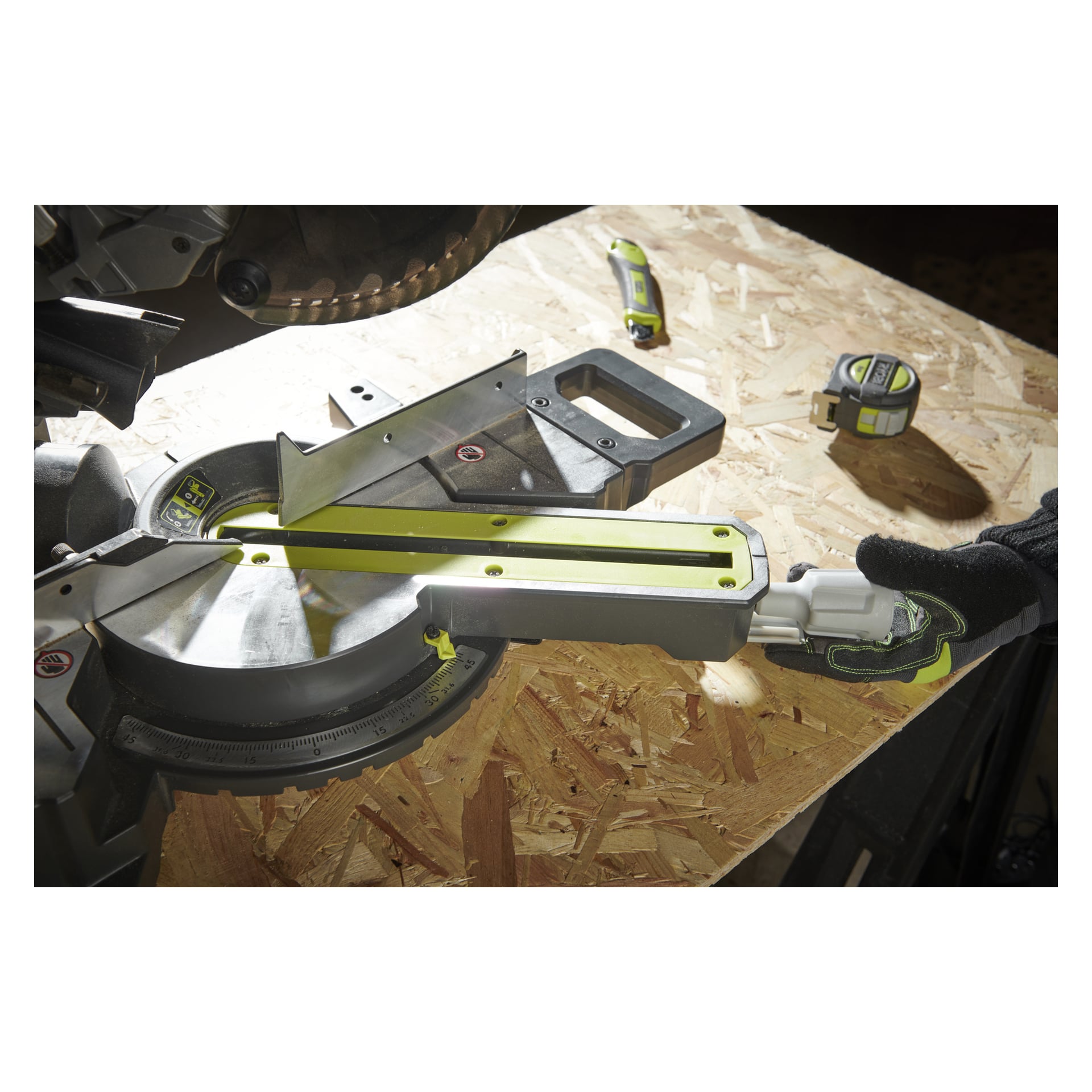 En detaljerad vy visar en hand med handske som justerar en Ryobi One+ batteridriven geringssåg RMS18190-0 placerad på en träyta, med ett måttband och en hobbykniv placerade längre bak på arbetsbänken.